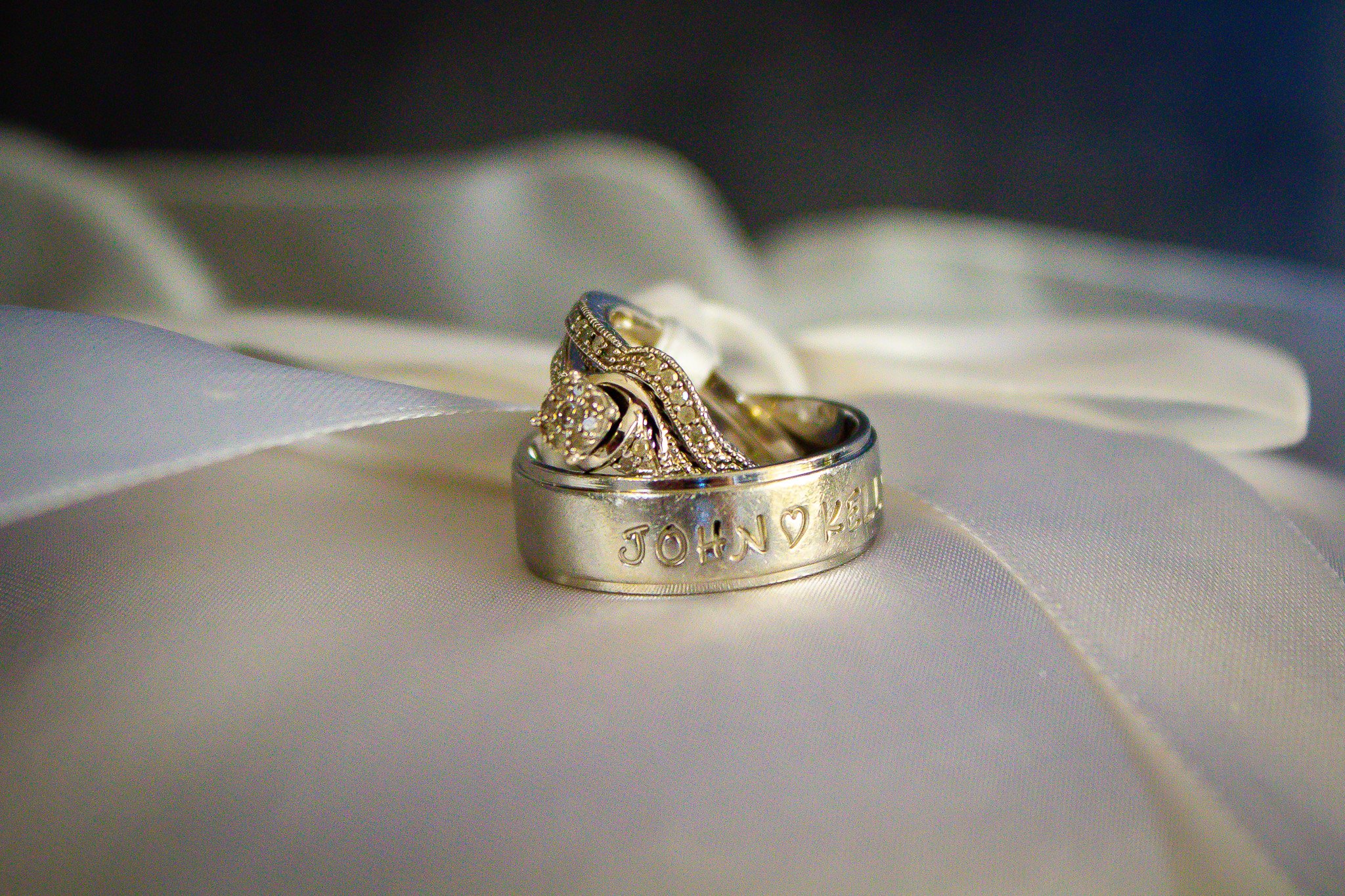 Close-up of a silver wedding band engraved with the names 'JOHN' and 'KELLY' with a heart symbol between, resting on white satin fabric with a white ribbon tied in a bow in the background.