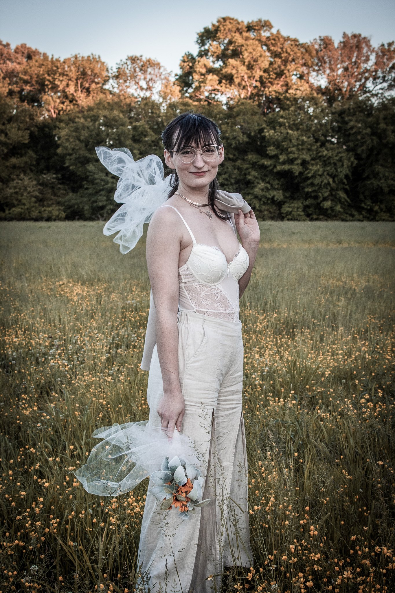A woman standing in a field of yellow flowers, holding a bouquet, wearing a white lace top and cream-colored pants, with a veil tied around her hair, during sunset.