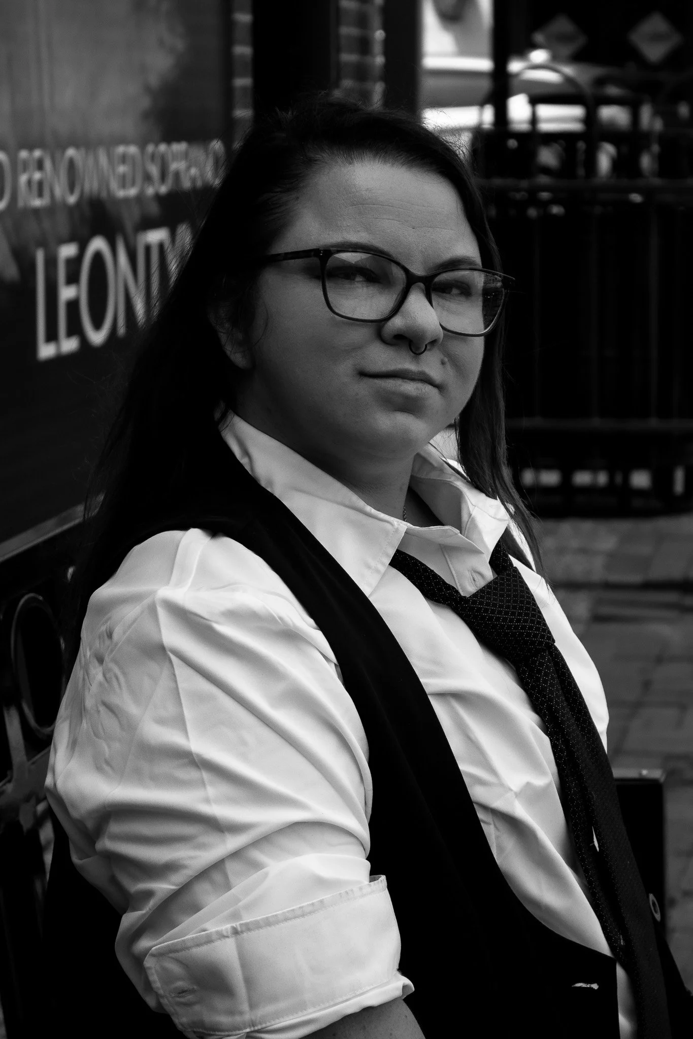 Black and white photo of a woman wearing glasses, a white shirt, a black vest, and a tie, standing outdoors.