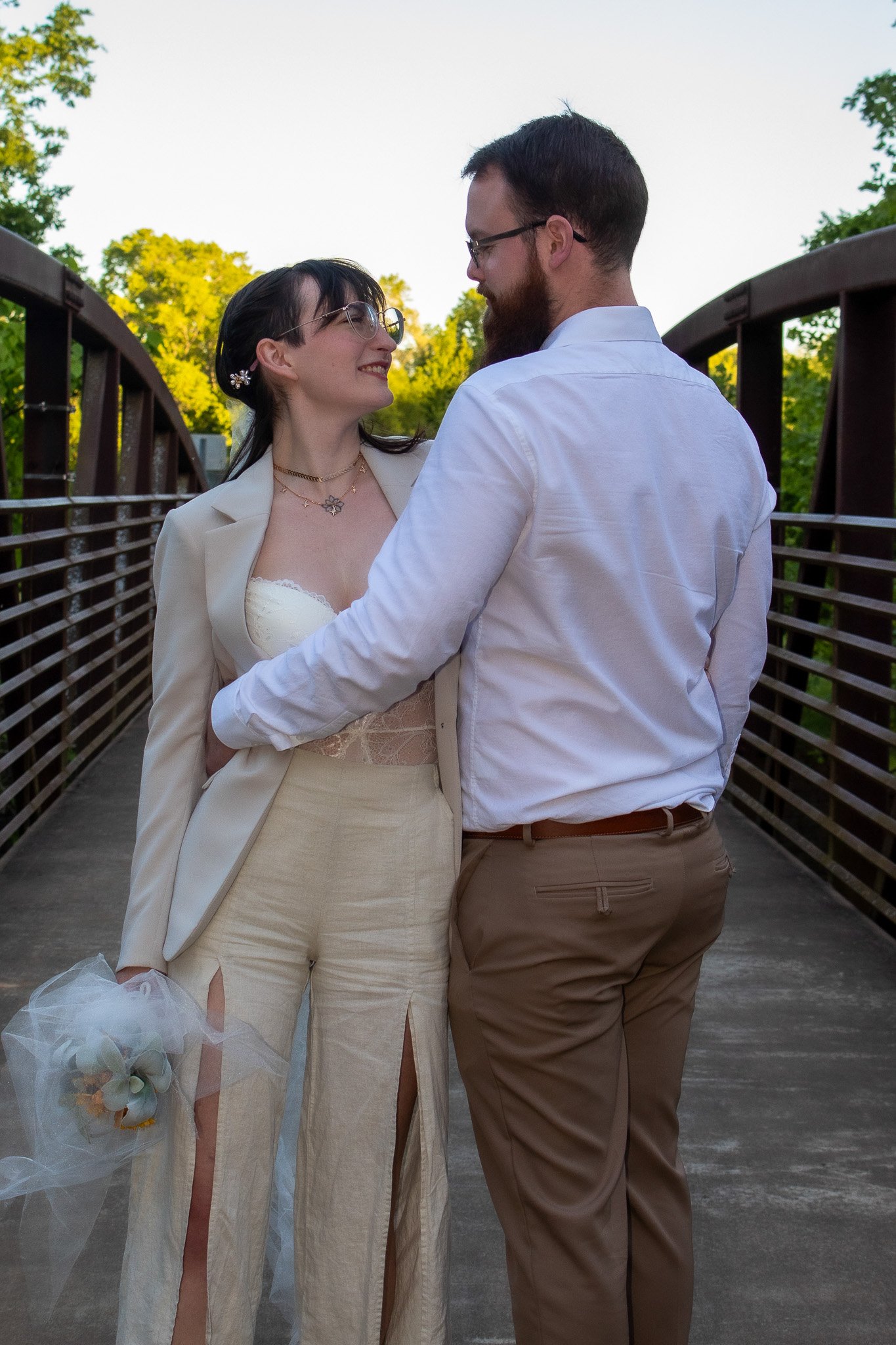 A couple holding Hands on a bridge during sunset, the woman is wearing a cream-colored blazer and pants with a satin top, holding a bridal bouquet. The man is dressed in a white shirt and khaki pants.