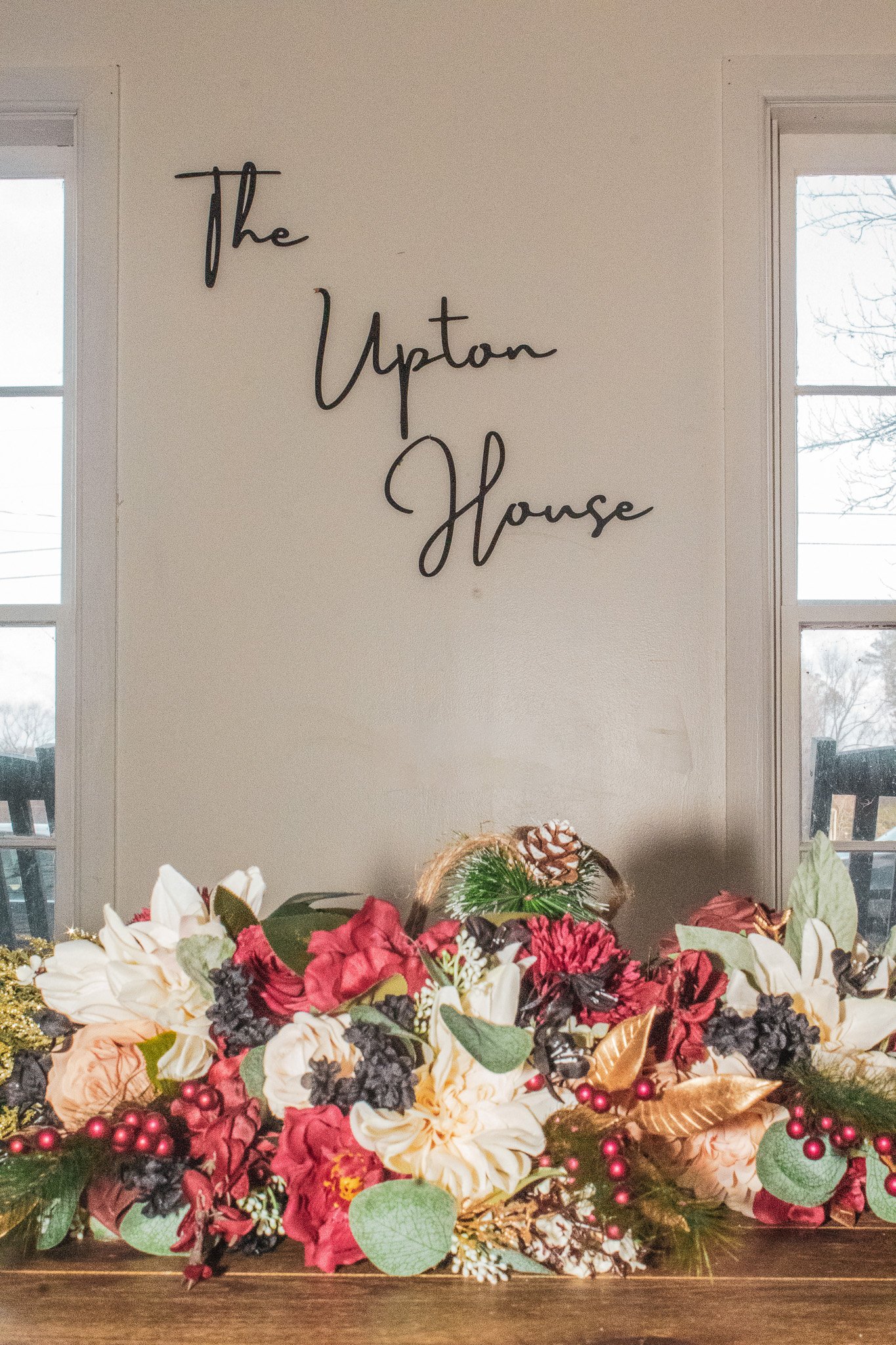 Decorative sign reading 'The Upton House' above a floral arrangement with red, white, and dark purple flowers, green leaves, and gold accents, on a wooden surface with windows in the background.