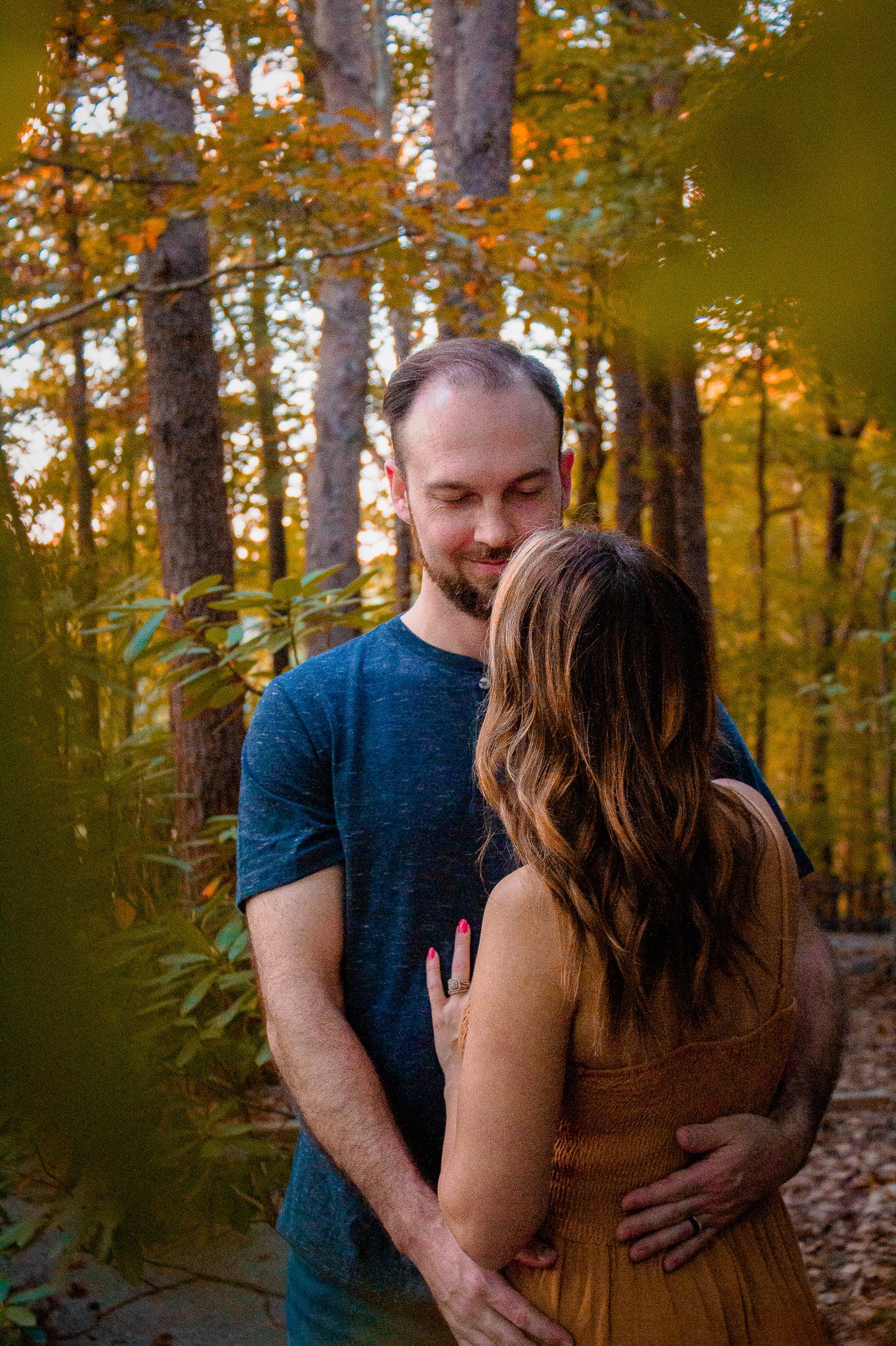 A man and woman embrace in a forest with autumn foliage, the man with closed eyes, the woman with long wavy hair, both smiling softly.