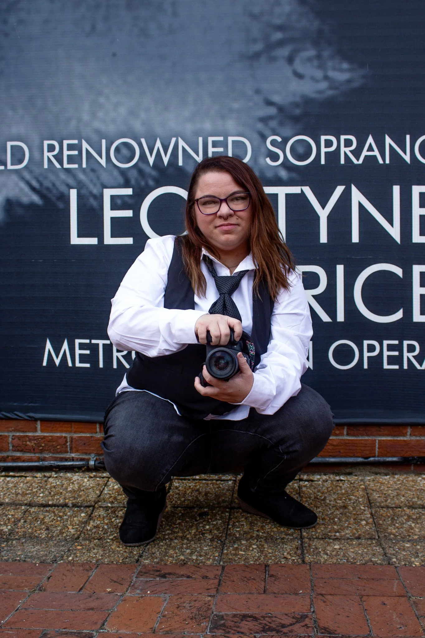 A person crouching outdoors in front of a large promotional poster, holding a camera and looking at the viewer, with long brown hair, glasses, a white shirt, black vest, and black pants.