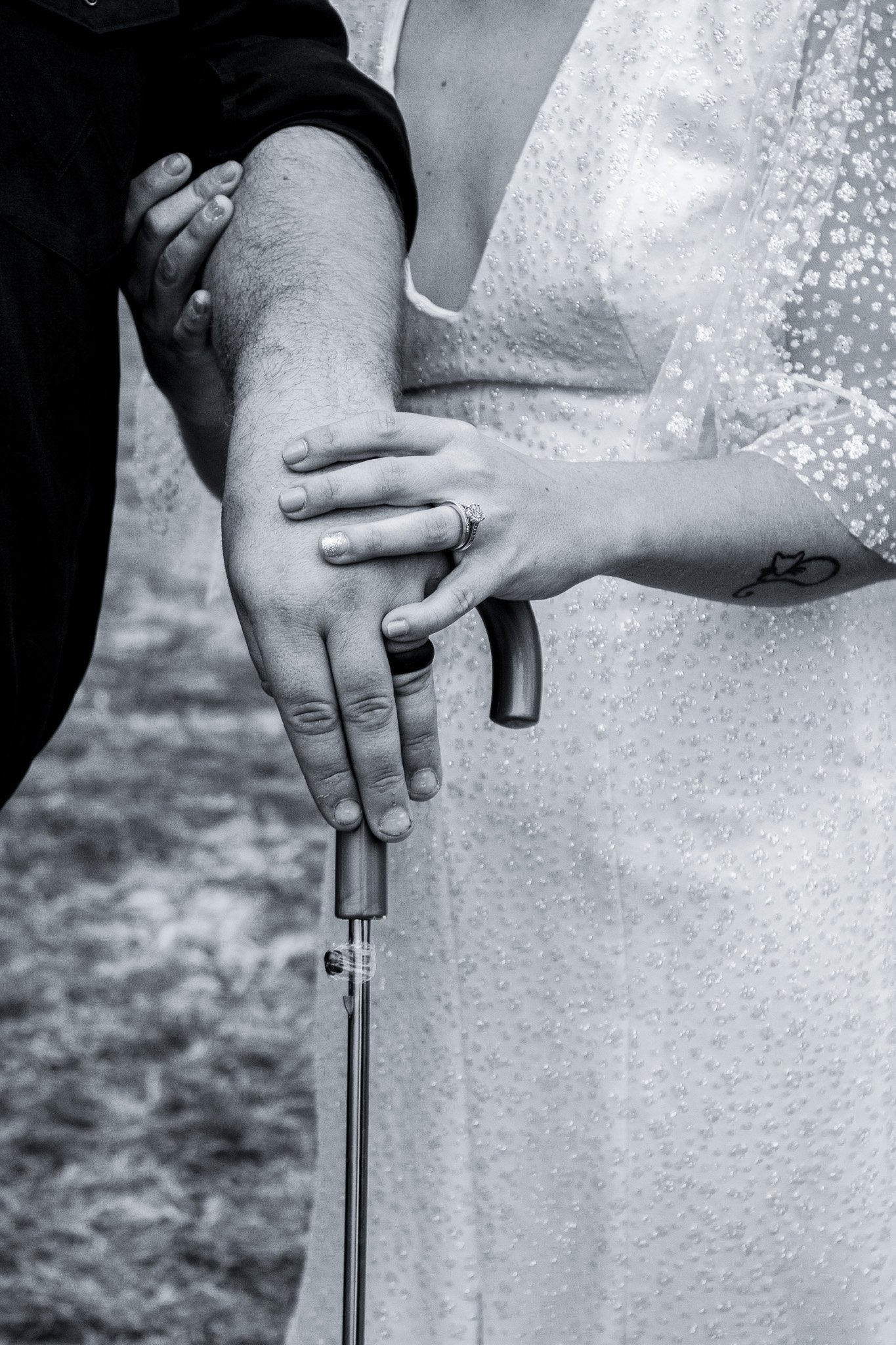 Close-up of two people holding a clear umbrella in the rain, with one person's hand resting on the other's arm. One person is wearing a ring on their finger, and both hands are grasping the umbrella handle.