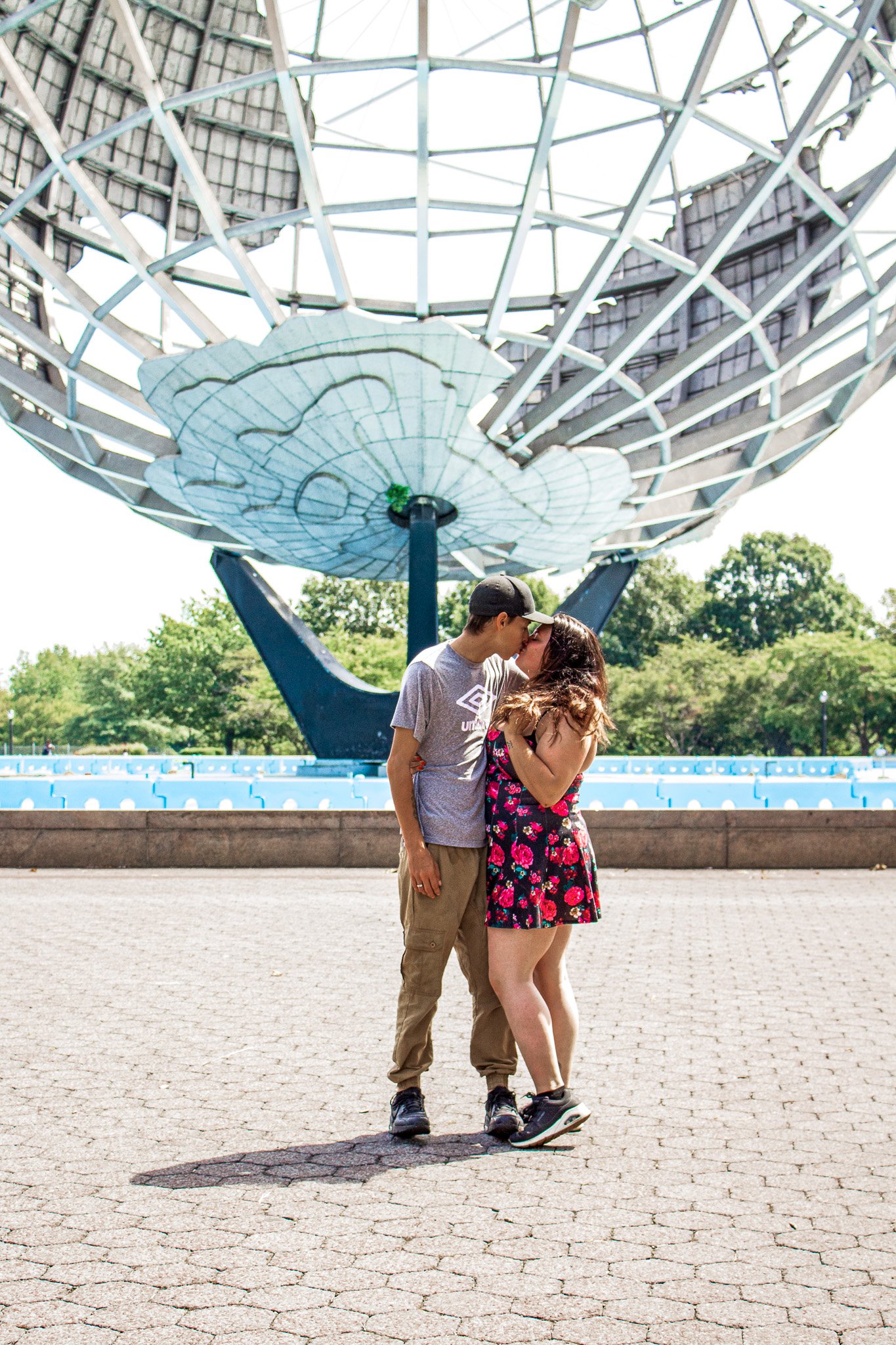 A young couple sharing a kiss in front of a large, spherical, metal and glass sculpture in an outdoor park area with green trees in the background.
