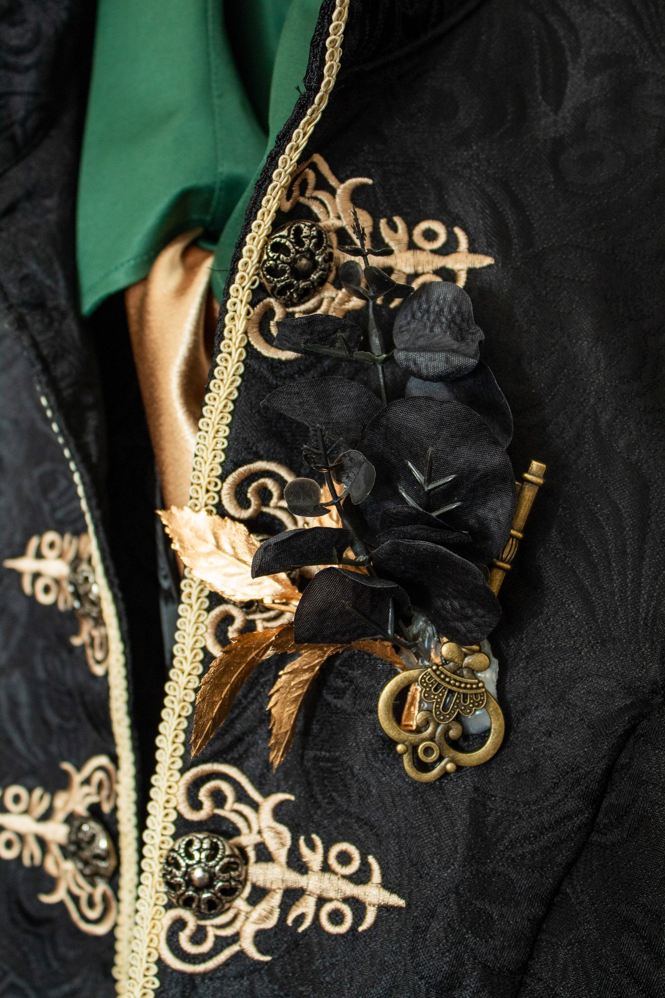 A black embroidered jacket with gold accents, decorative buttons, and a floral brooch with black and gold leaves, attached to the jacket.