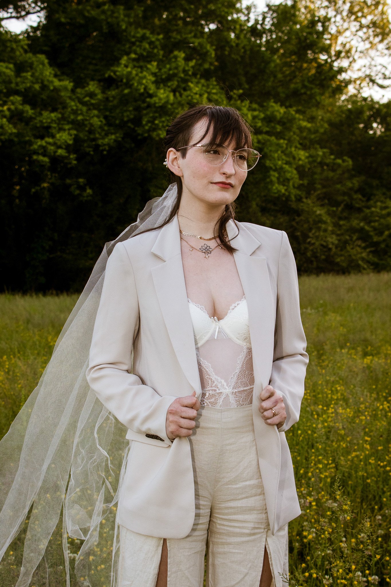 A woman in a cream-colored suit with lace lingerie underneath, standing outdoors in a field of yellow flowers and green trees, wearing glasses and a veil.
