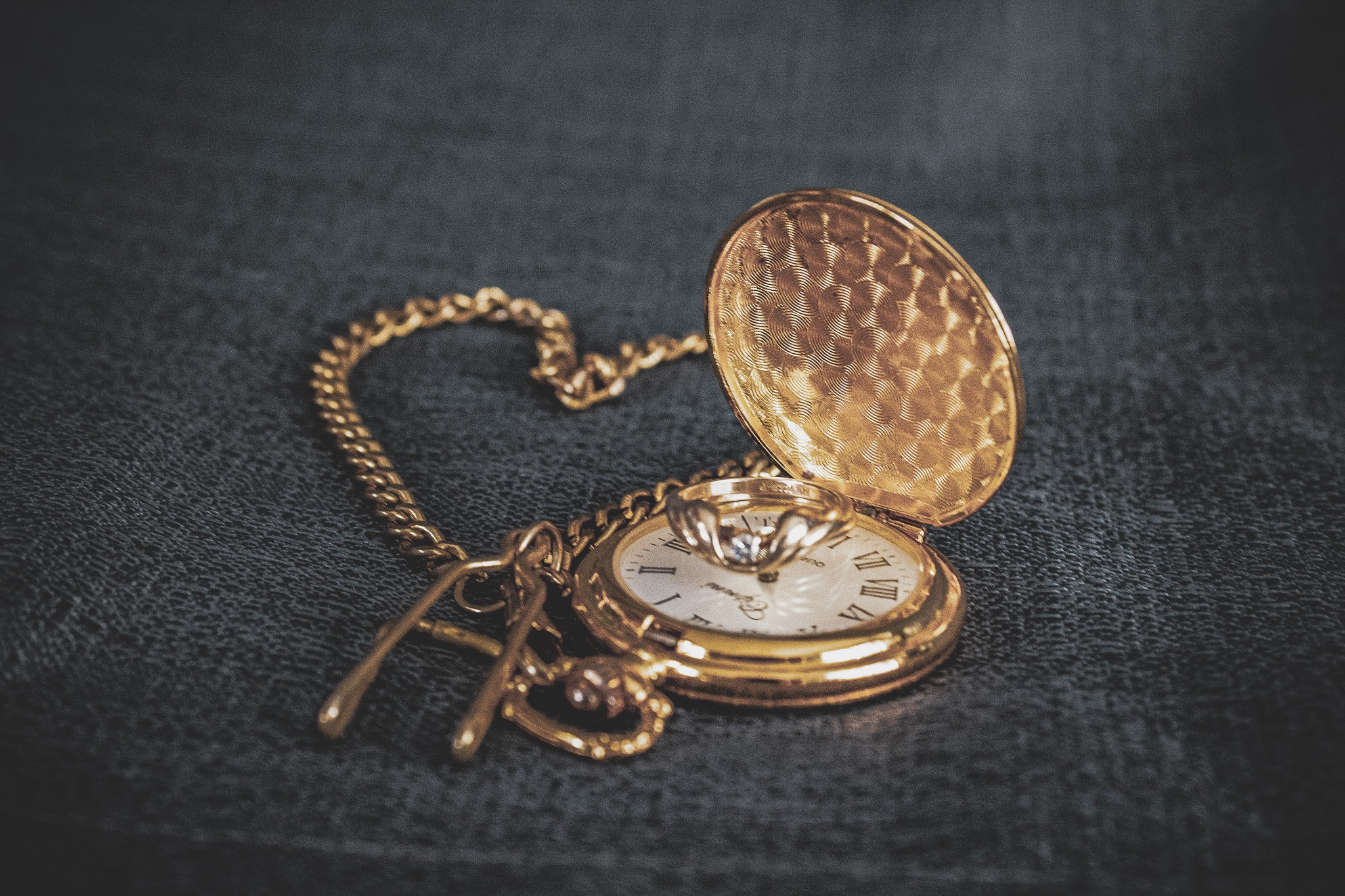 Gold pocket watch with open cover and chain, lying on black textured fabric.