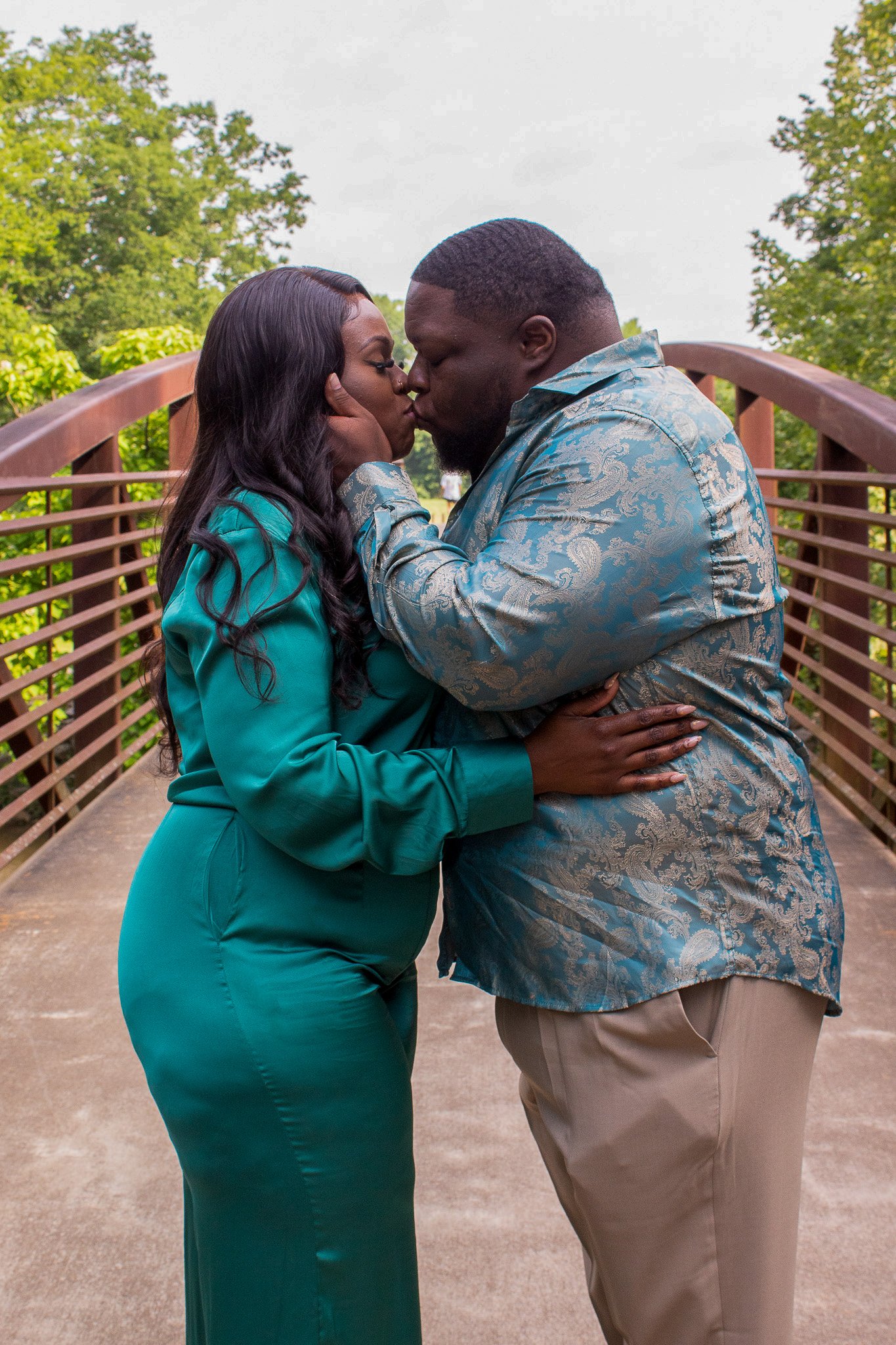 A couple sharing a kiss on a bridge, surrounded by green trees, with the woman in a teal dress and the man in a patterned blue shirt.