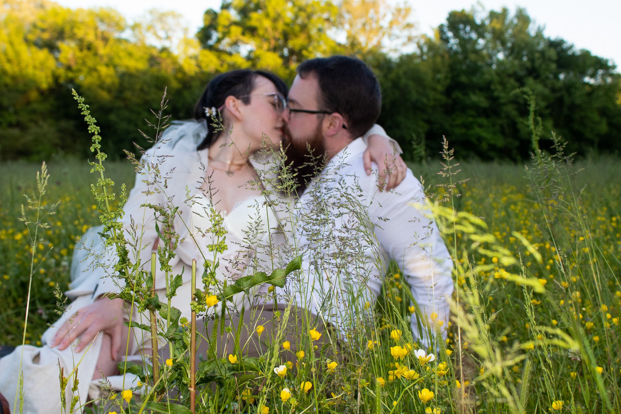A couple kissing in a field of tall grass and yellow flowers, with green trees and a clear sky in the background.