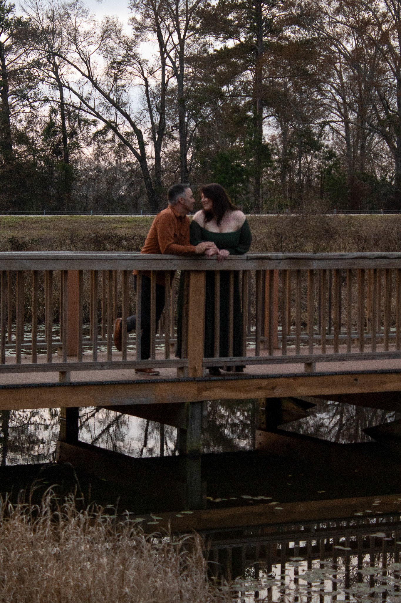 A couple stands on a wooden bridge over a pond, looking at each other lovingly, with leafless trees in the background during dusk.