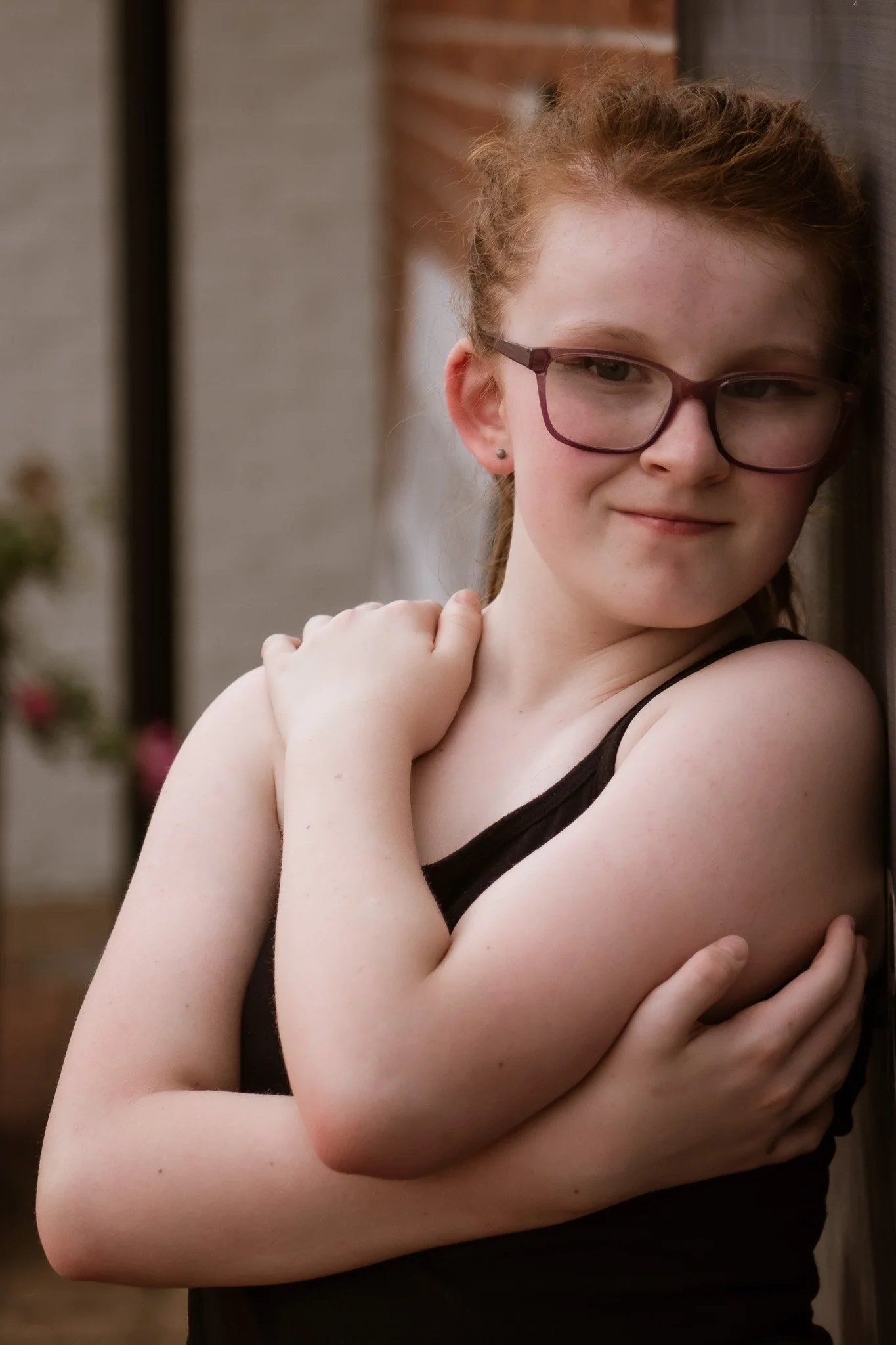A young girl with red curly hair, wearing glasses and a black sleeveless top, leaning against a wooden wall outdoors with a gentle smile.