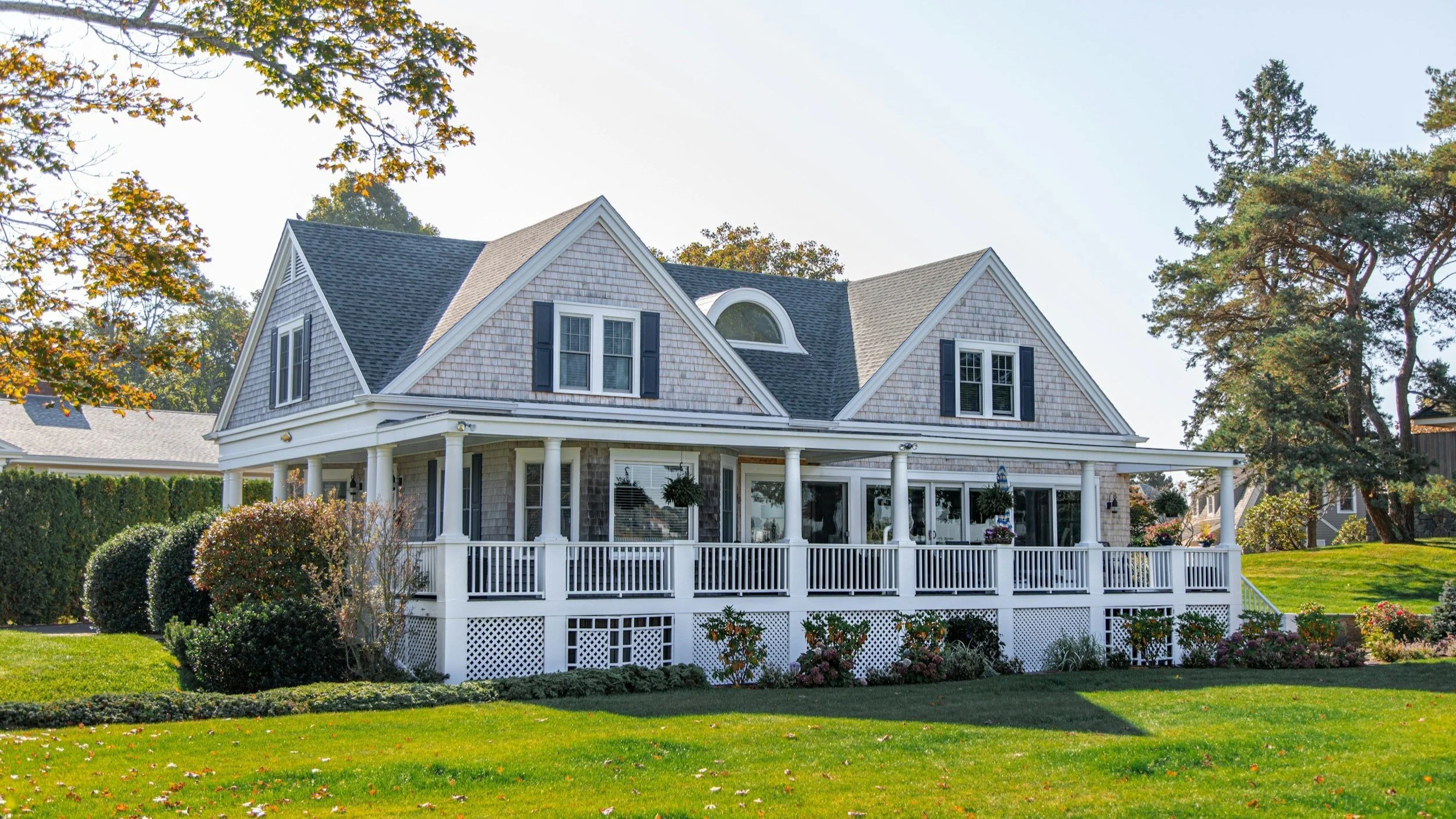 A large house with a gray shingle exterior, black shutters, and a spacious white porch, surrounded by a well-maintained lawn and trees, in a suburban neighborhood.