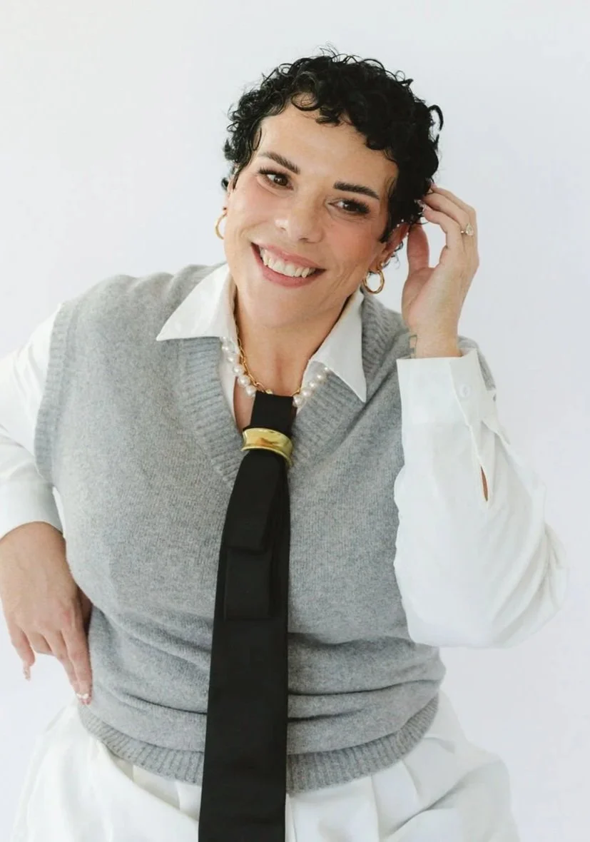 A woman with short curly black hair, hoop earrings, and a gold name necklace smiling at the camera against a plain light background.