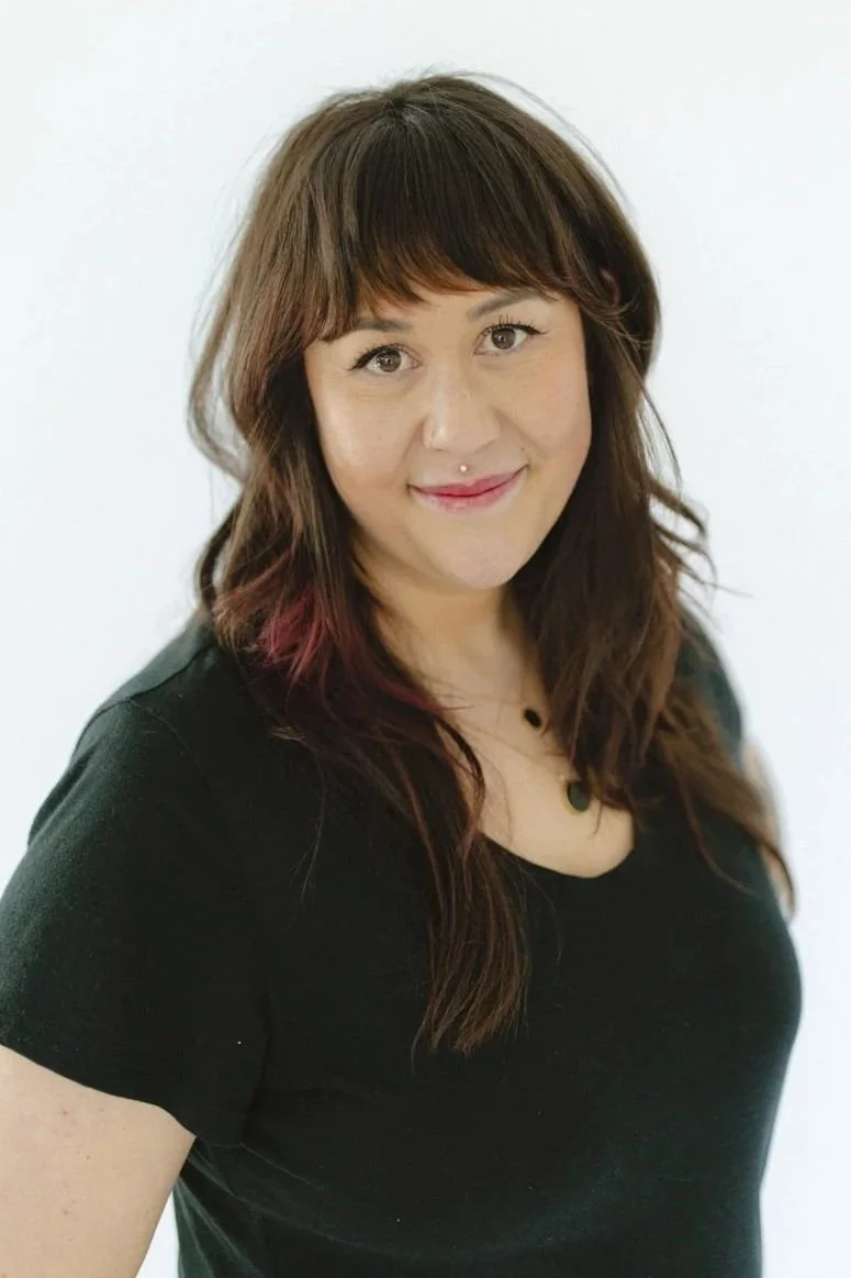 A woman with long dark hair and a sleeve tattoo on her right arm stands indoors, smiling at the camera.