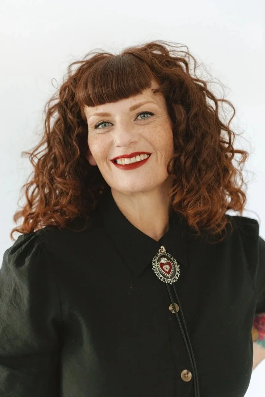 A confident woman with red hair styled in voluminous curls and blunt bangs, wearing a black button-up dress with puffed sleeves. She has layered necklaces, a large statement ring, and multiple bracelets. Her makeup includes red lipstick, and her right hand, which is on her hip, reveals a floral tattoo.