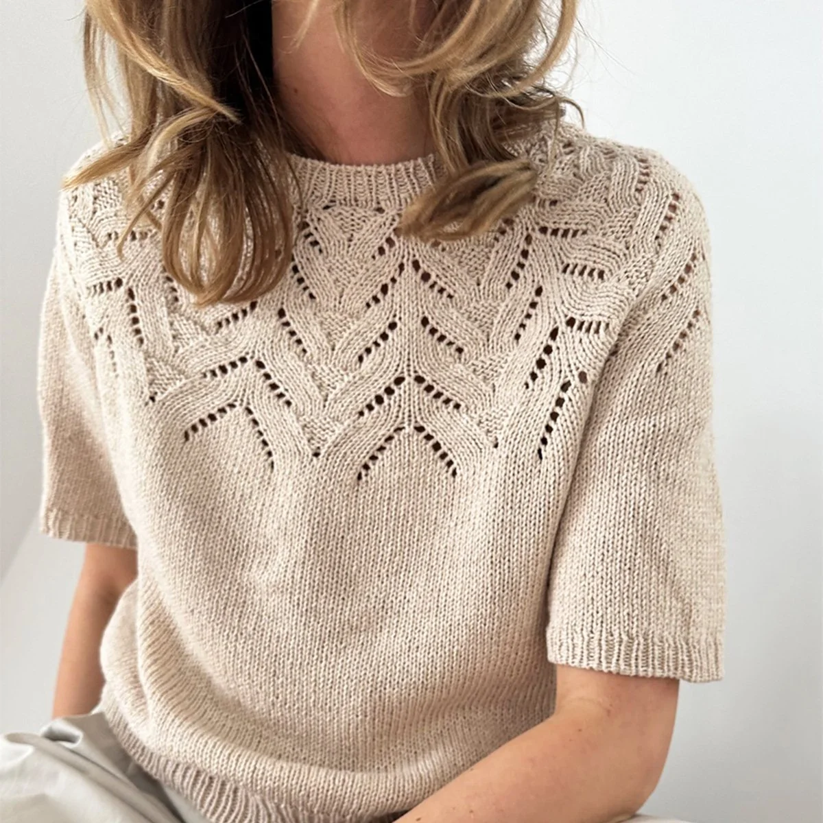 A woman wearing a beige knitted sweater with lace-like patterns on the chest and shoulders, and short sleeves.