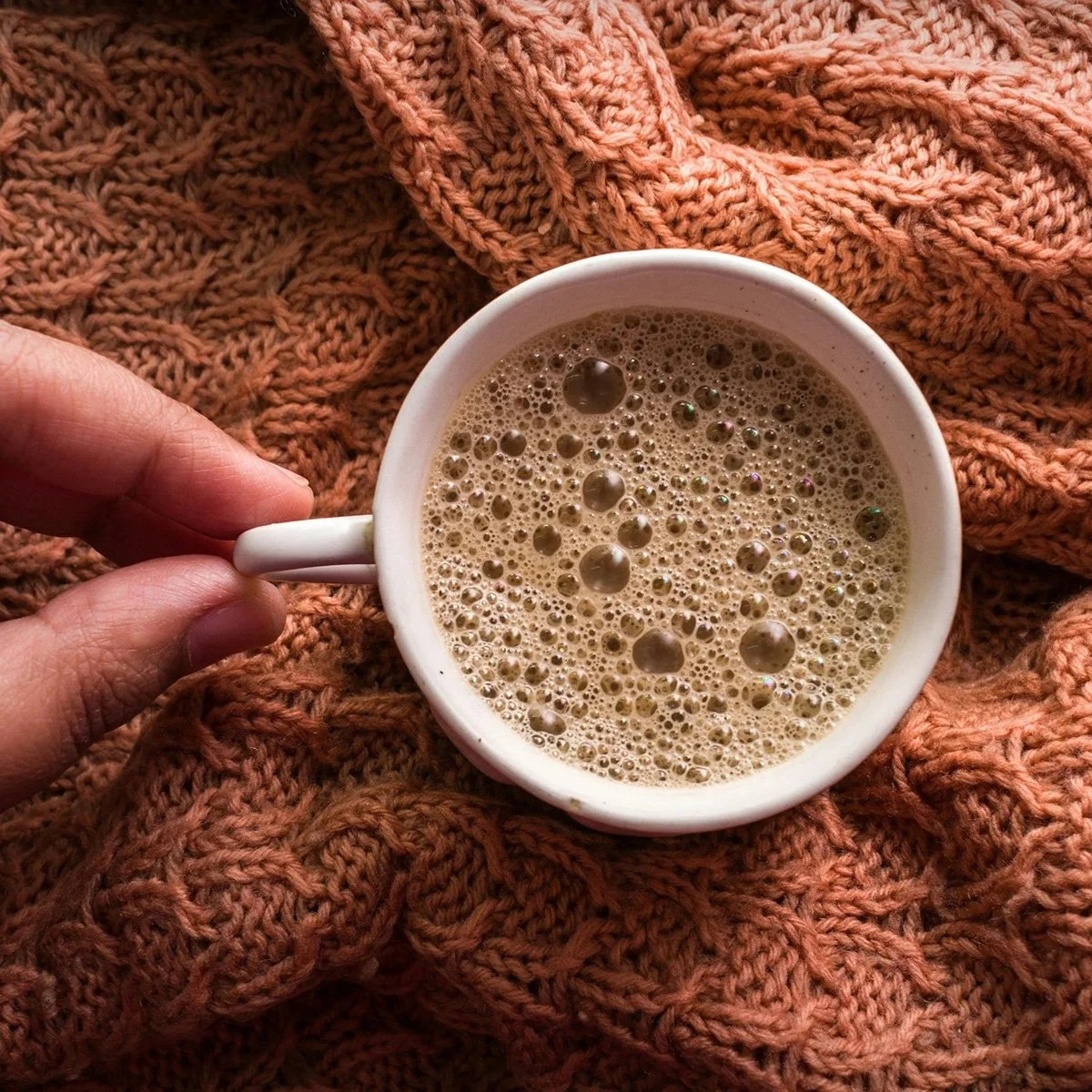 A white mug filled with frothy coffee held by a person's hand, placed on a textured orange knitted blanket.