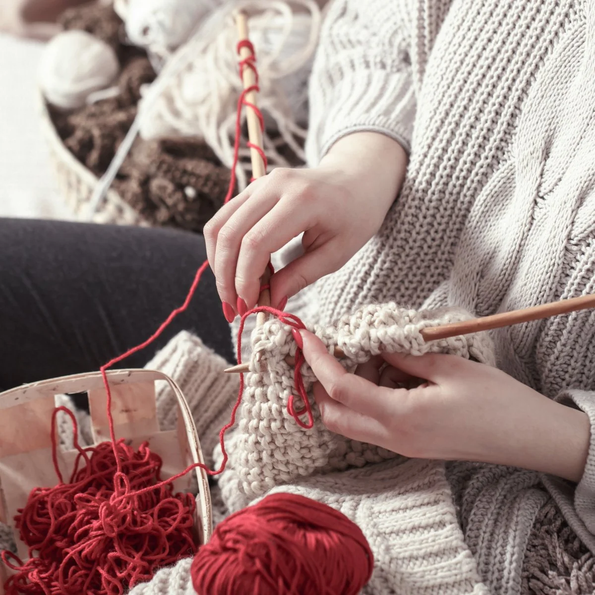 Person knitting with white yarn, surrounded by red yarn balls, using knitting needles, with a textured beige sweater.