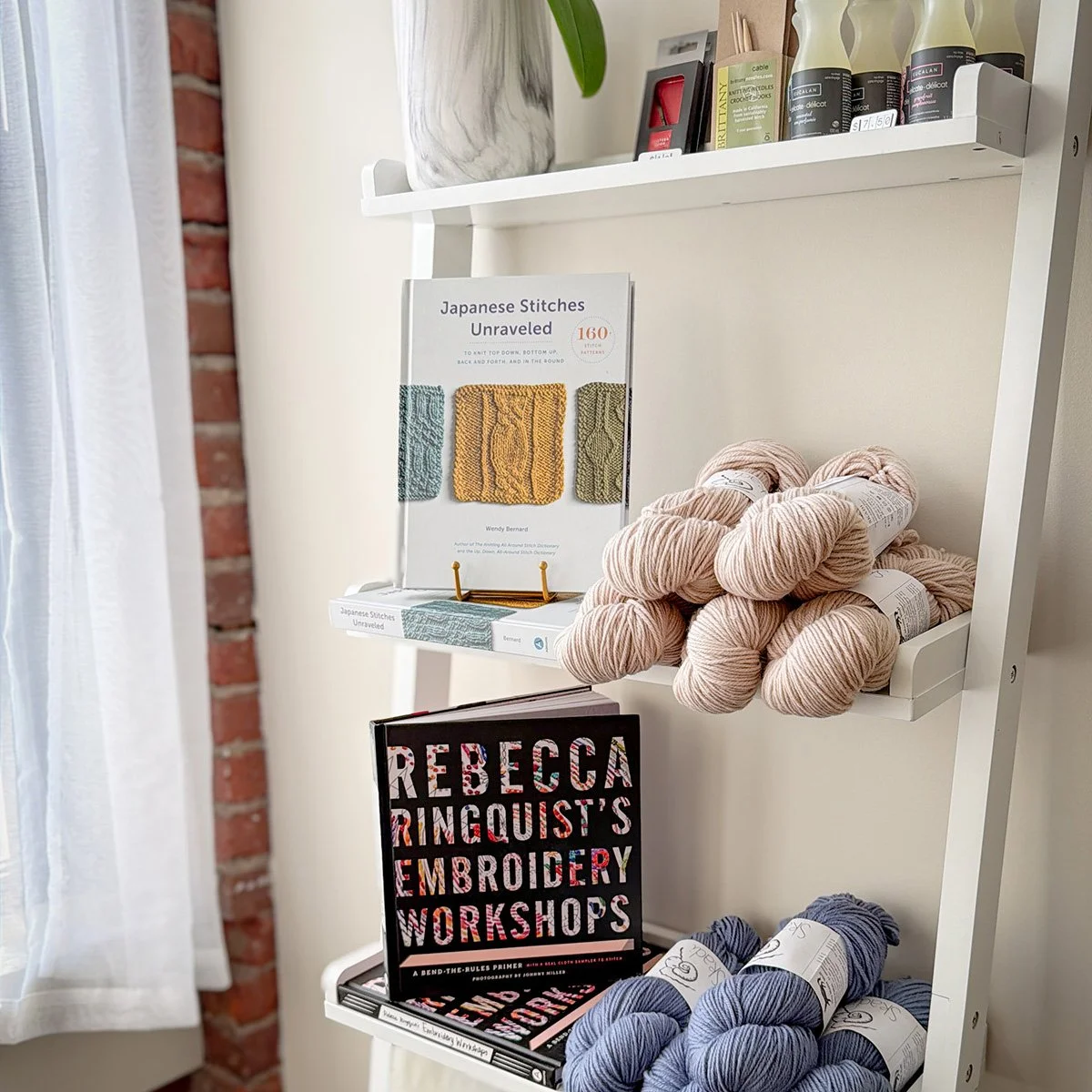 Shelf with knitting and embroidery books, yarn skeins in pink and blue, and a plant in a white pot against a white wall with a brick wall nearby.