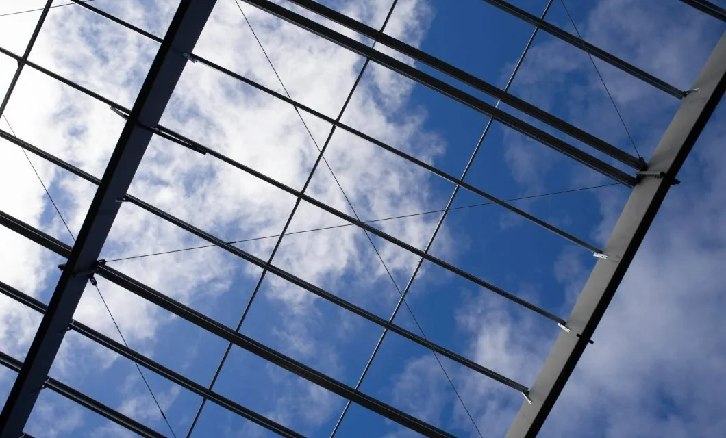 Looking up at a glass ceiling with a metal framework against a partly cloudy blue sky.