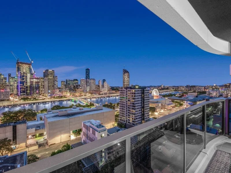 City skyline at dusk viewed from a high-rise balcony with glass railing, featuring illuminated buildings and a river in the background.