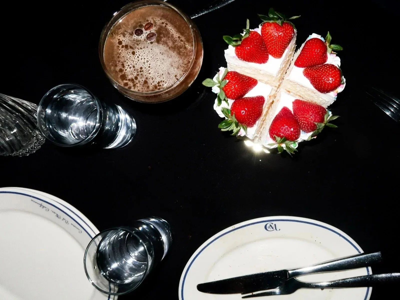 A slice of strawberry cake on a black table, with a glass of chocolate beverage, water glasses, white plates, and forks nearby.