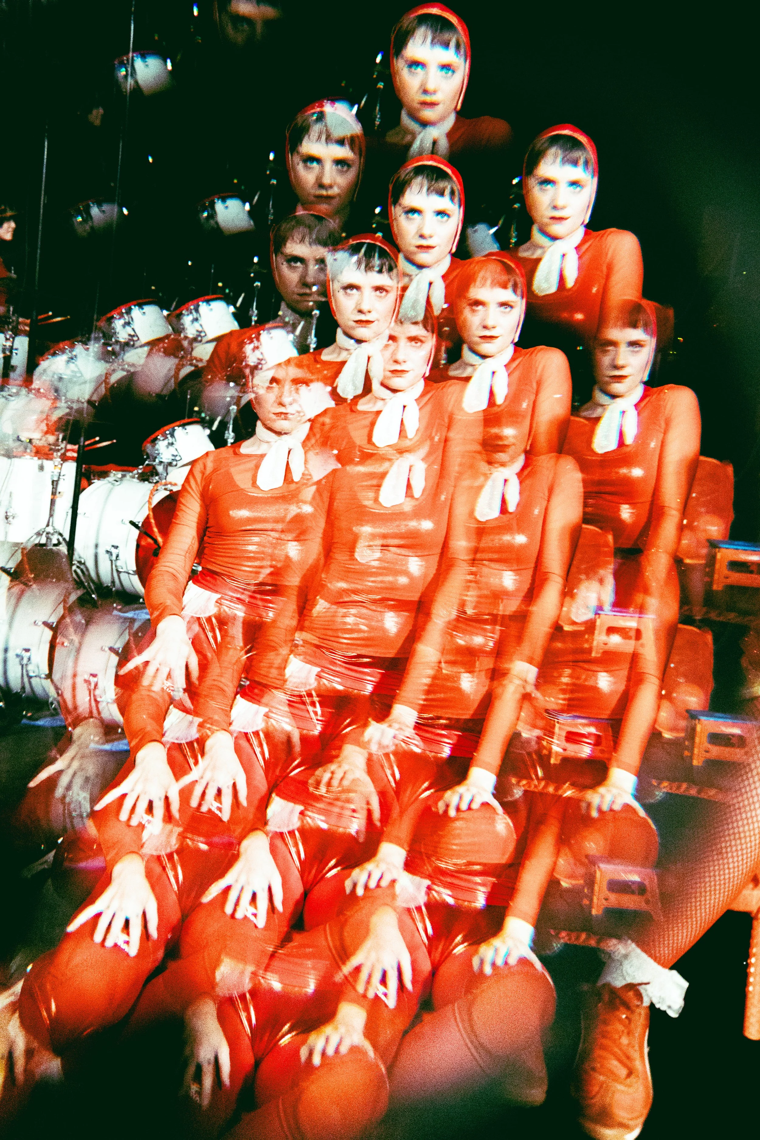 A multi-exposure image of a woman dressed in a red outfit with a white bow, sitting on red suitcases, with her reflection repeating multiple times in a mirrored background. GELLI HAHA LIVE AT THE ROXY.