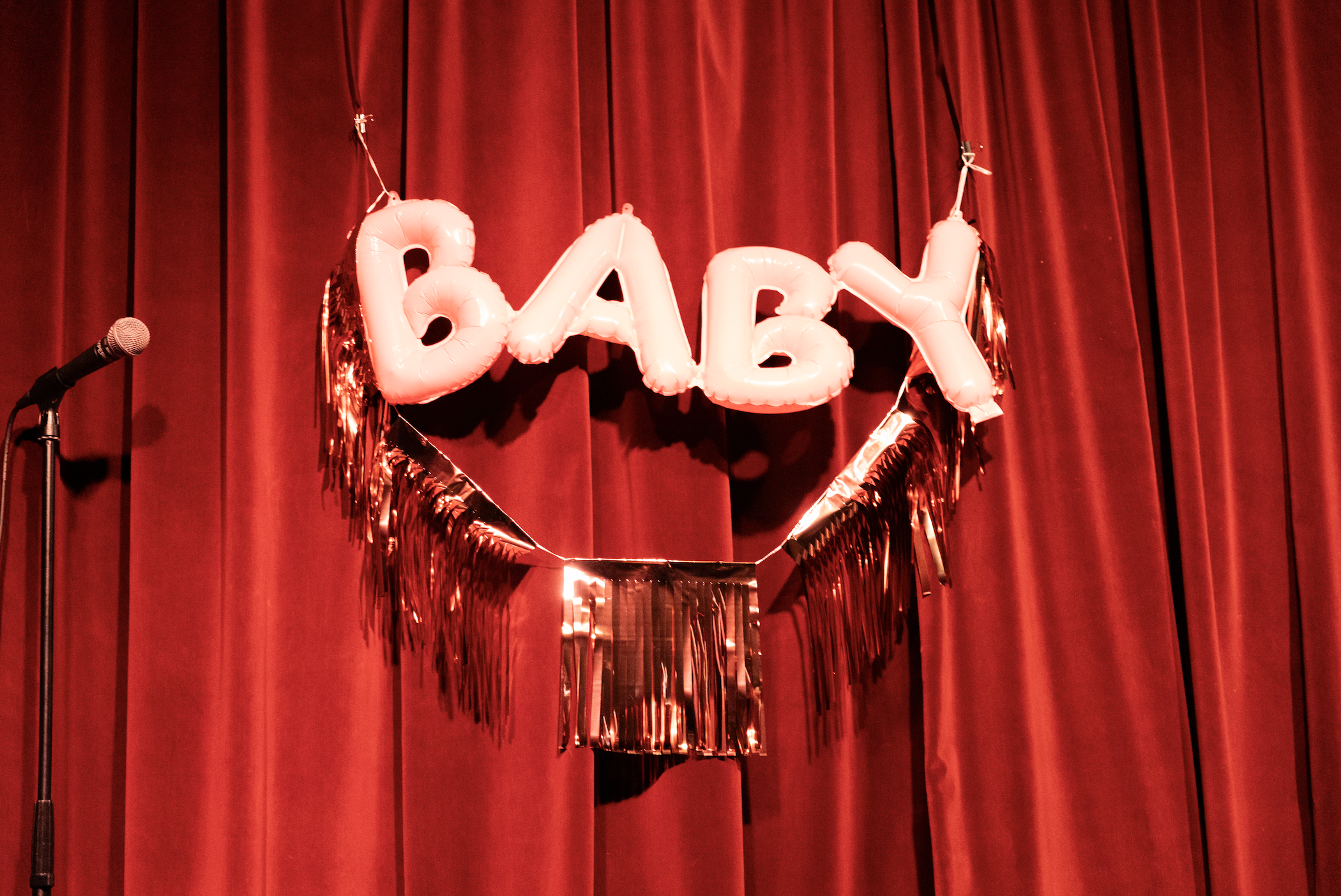 Decorative sign spelling 'BABY' in pink balloons, hanging in front of red stage curtains with metallic gold fringe accents.