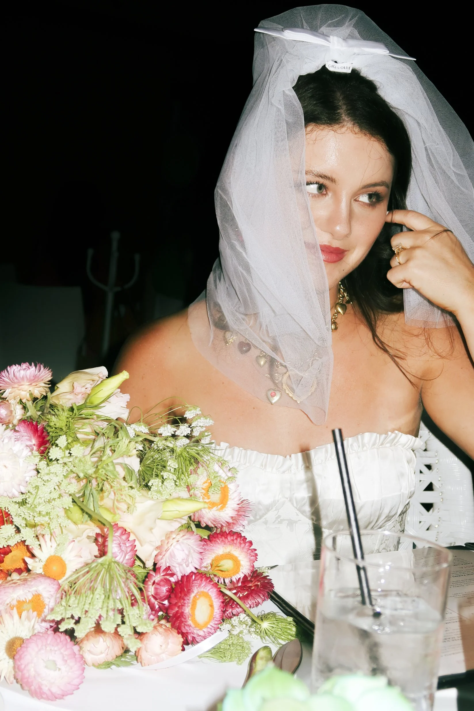 A woman in a white strapless dress wears a veil with a label reading "GIGI & OLIVE," sitting at a table with a colorful flower arrangement, drinking with a black straw, and touching her face with her right hand.