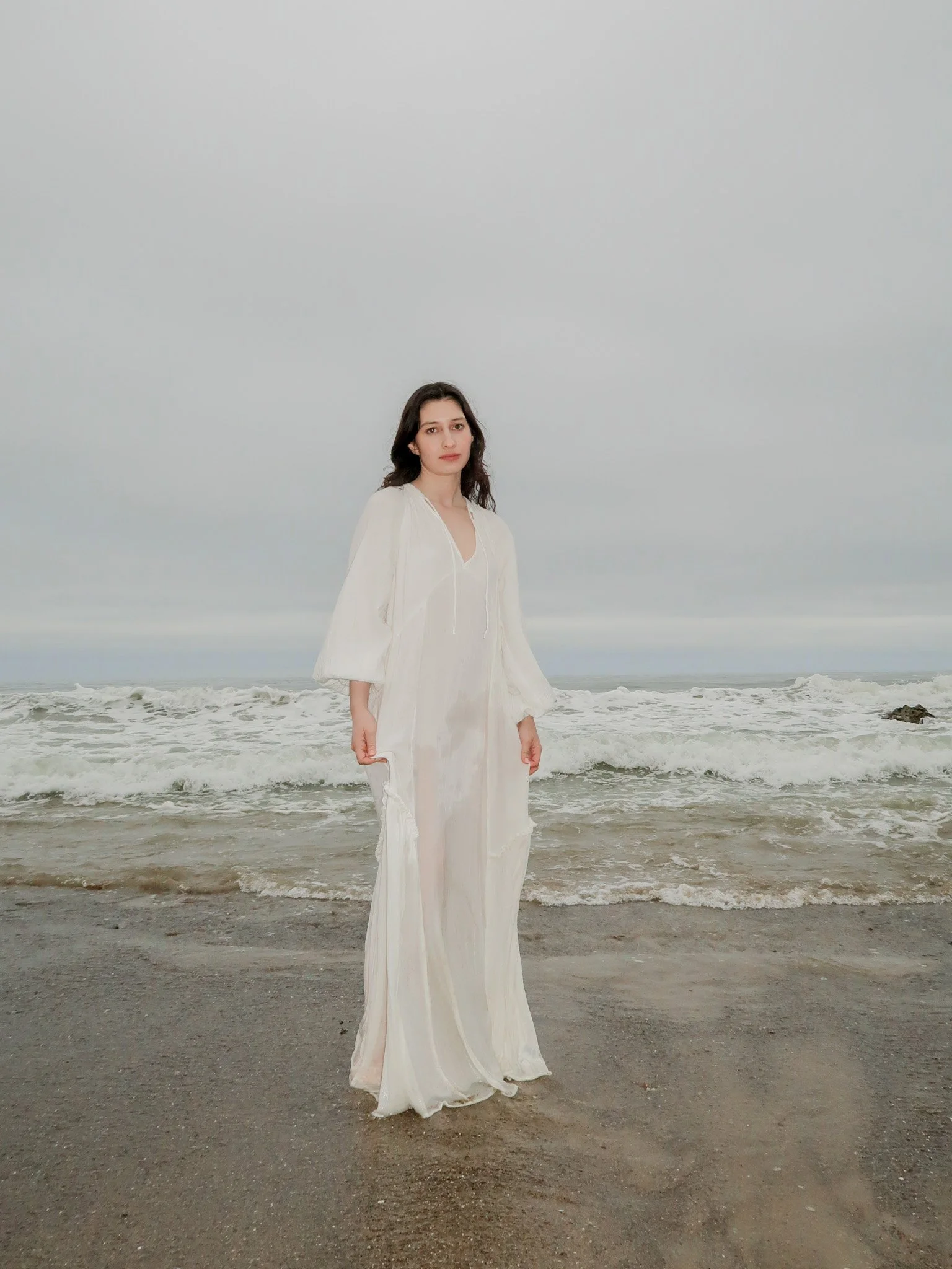 A woman in a flowing white dress stands on a sandy beach near the ocean, with waves and cloudy sky in the background.