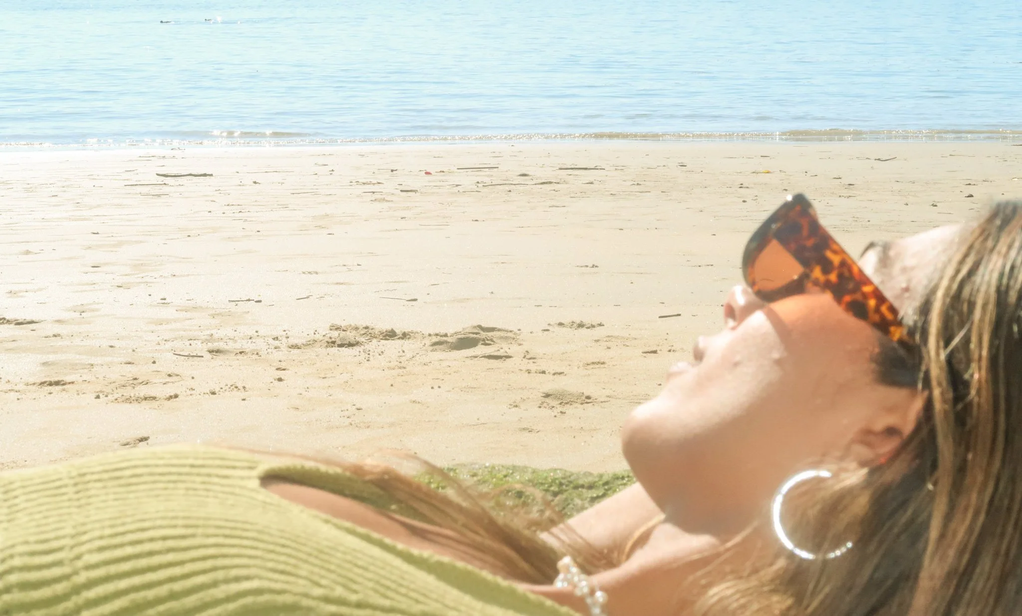 A woman with long hair, wearing tortoiseshell sunglasses, hoop earrings, and a necklace, relaxing on the beach with her eyes closed as she basks in the sun.
