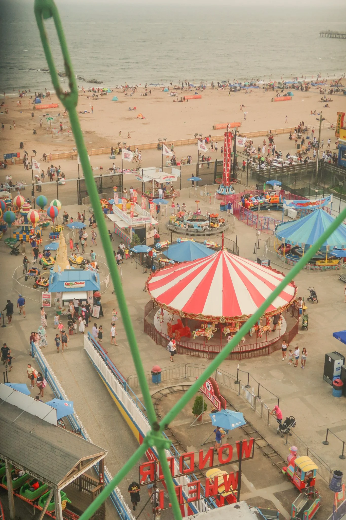 A lively amusement park near a beach with colorful rides, a carousel, and many visitors, seen from an elevated vantage point through a green wire fence.
