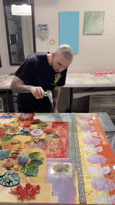 An artist uses a heat gun on a colorful mixed media art piece that includes flowers, a woman's face, and various textured elements, in a studio with artwork on the walls.