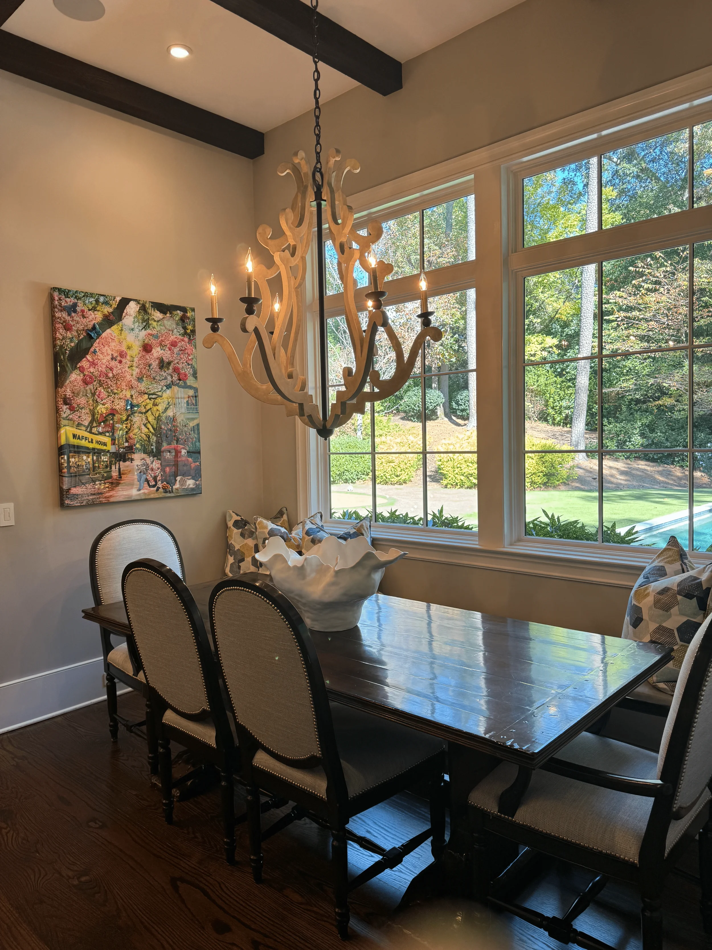Dining room with a dark wooden table, six beige upholstered chairs with black trim, a white decorative bowl, large windows showing a backyard with trees and grass, a beige wall with a colorful painting, and a chandelier with multiple candle-shaped lights hanging from the ceiling.