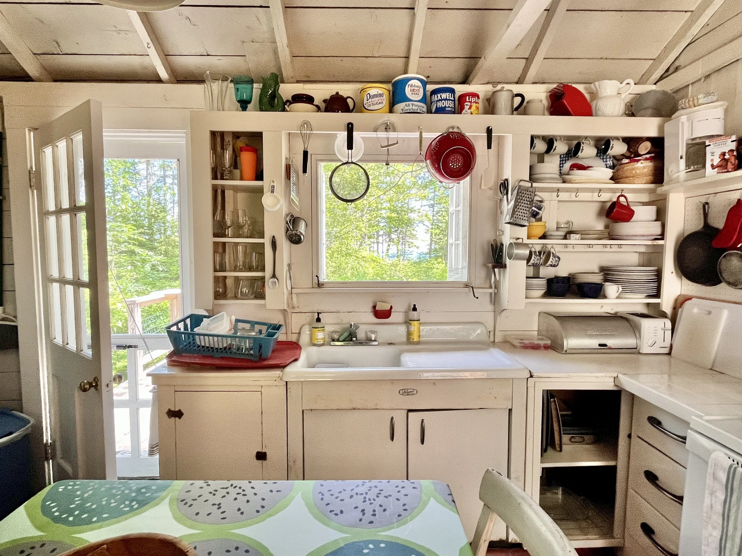 Cozy kitchen with open door, window, white cabinets, open shelves with dishes, hanging utensils, and a table with a colorful tablecloth.