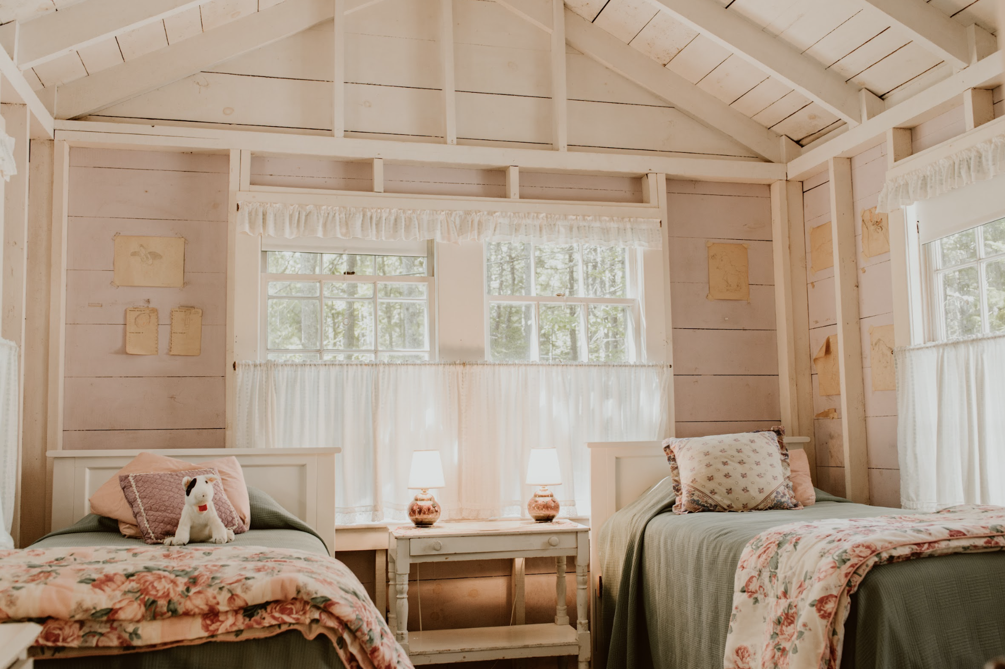 A cozy bedroom with two single beds, a white nightstand with two lamps, and windows with curtains. One bed has a pink pillow and a stuffed dog toy, while the other has patterned pillows. The room has light wood paneling and a high, peaked ceiling.