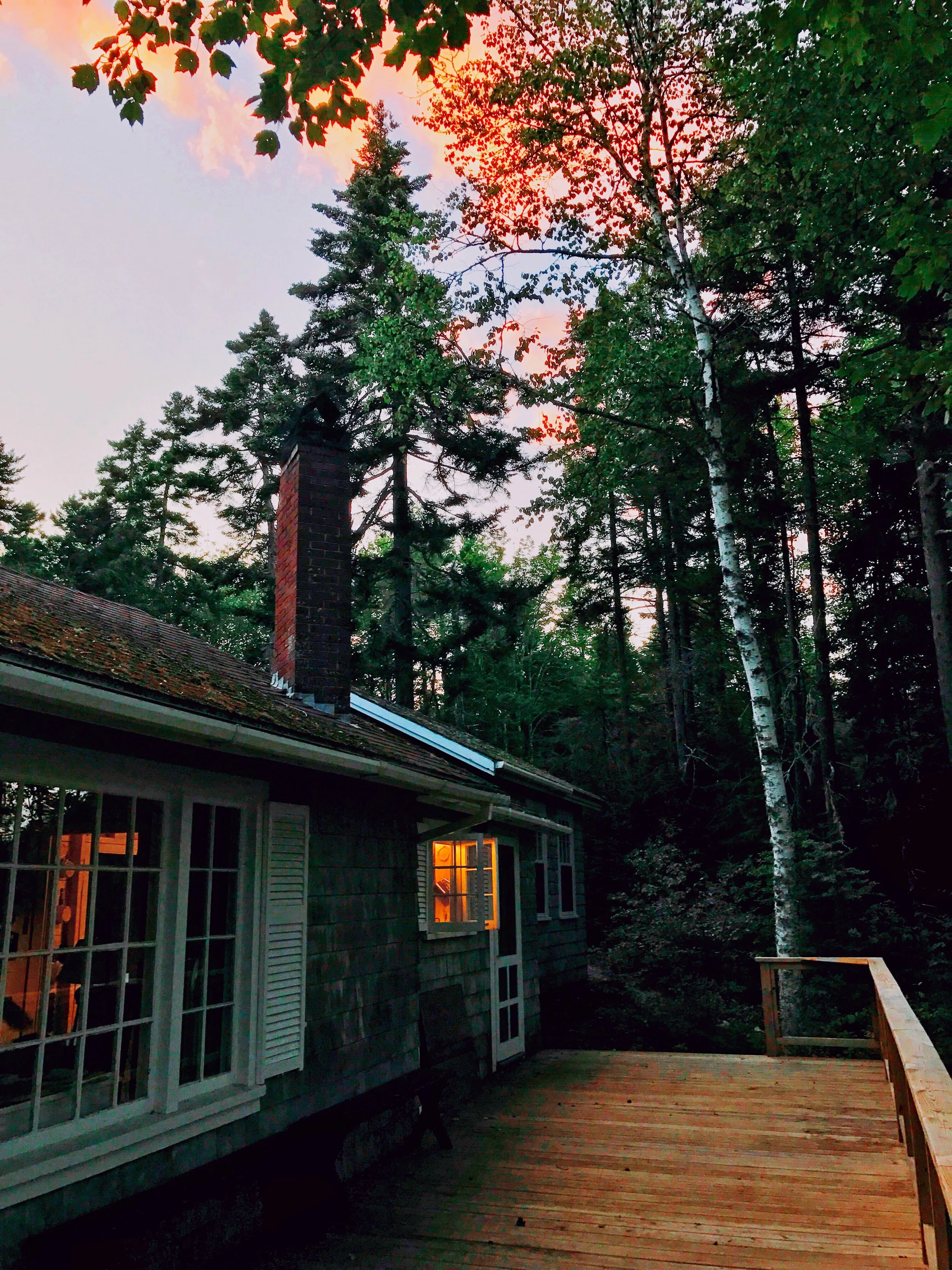 A wooden deck attached to a house with a chimney, surrounded by tall trees, during sunset with colorful sky.
