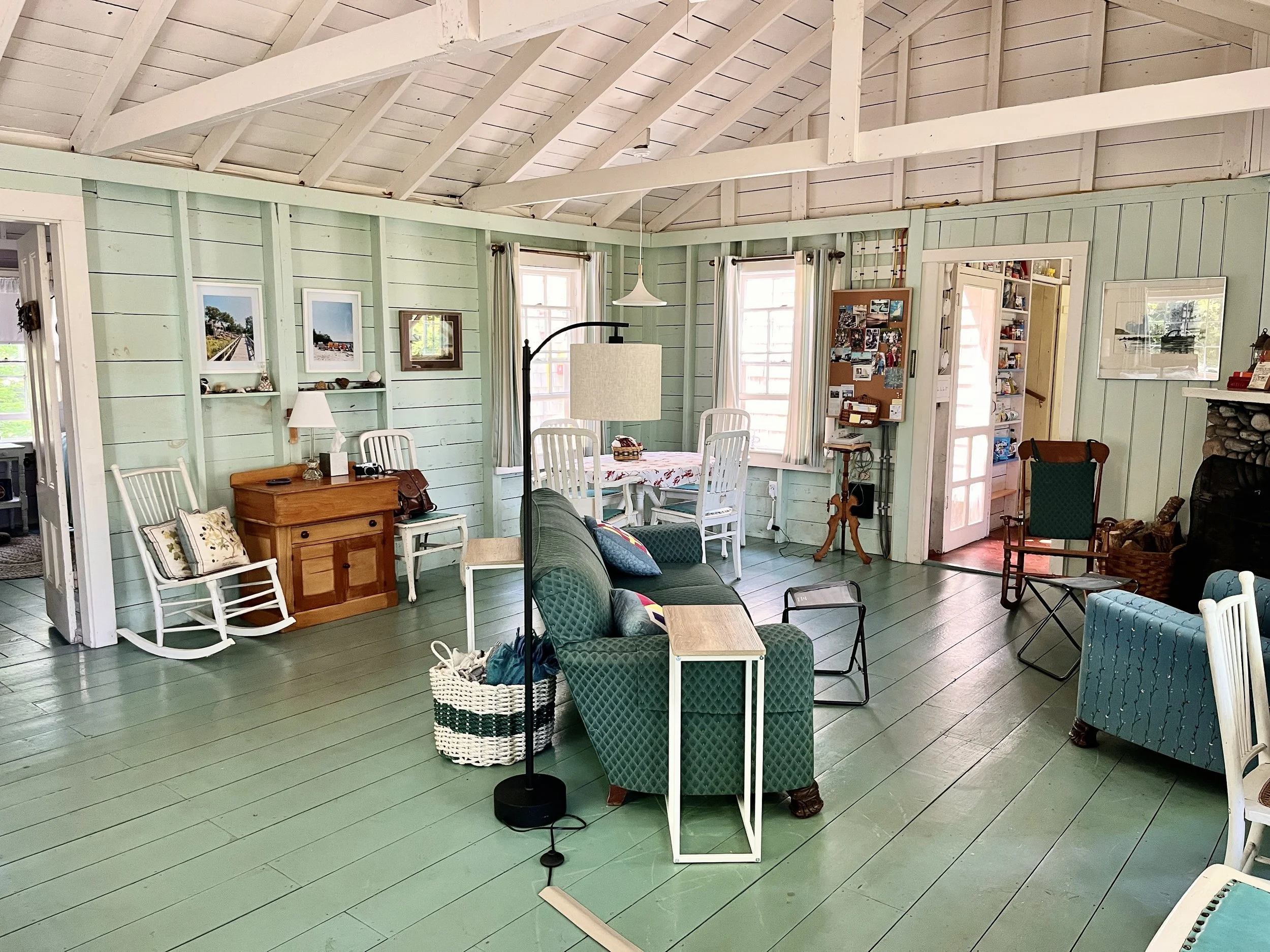 Living room with mint green wooden walls, white ceiling beams, and green wooden plank flooring. Contains a green upholstered armchair, a small black side table, a white rocking chair, a wooden cabinet, a dining table with white chairs, and various fr