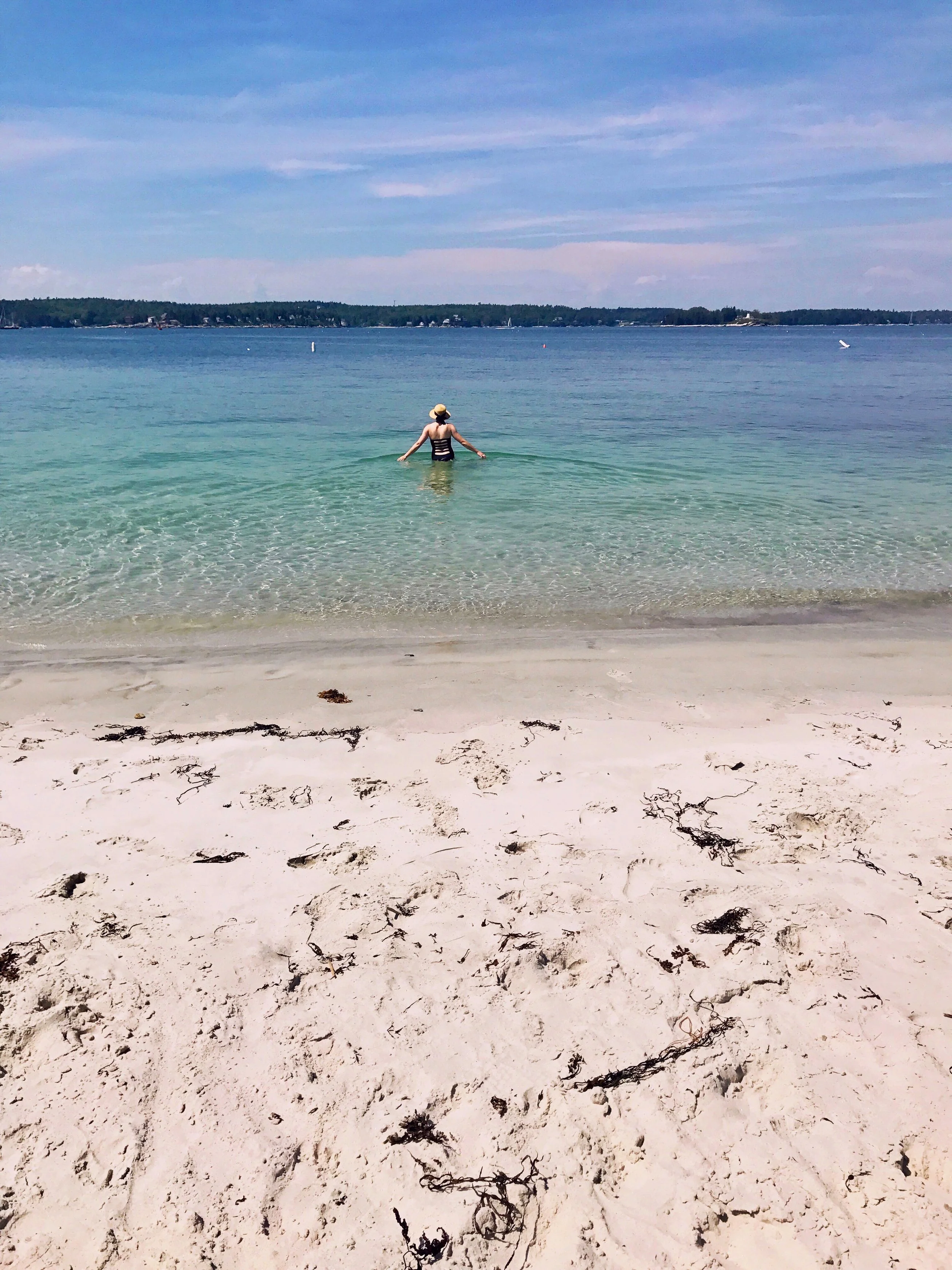 Person in a striped swimsuit and wide-brim hat standing in calm, clear water at the beach, with sandy shore and distant shoreline visible under a partly cloudy sky.