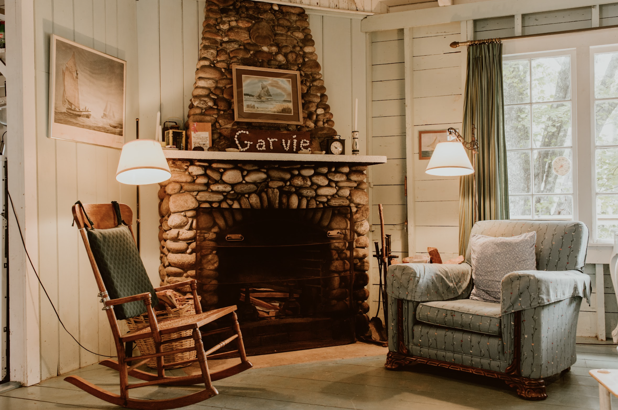 Living room with a stone fireplace, two lamps, a wooden rocking chair, an upholstered armchair, and framed pictures on the walls.