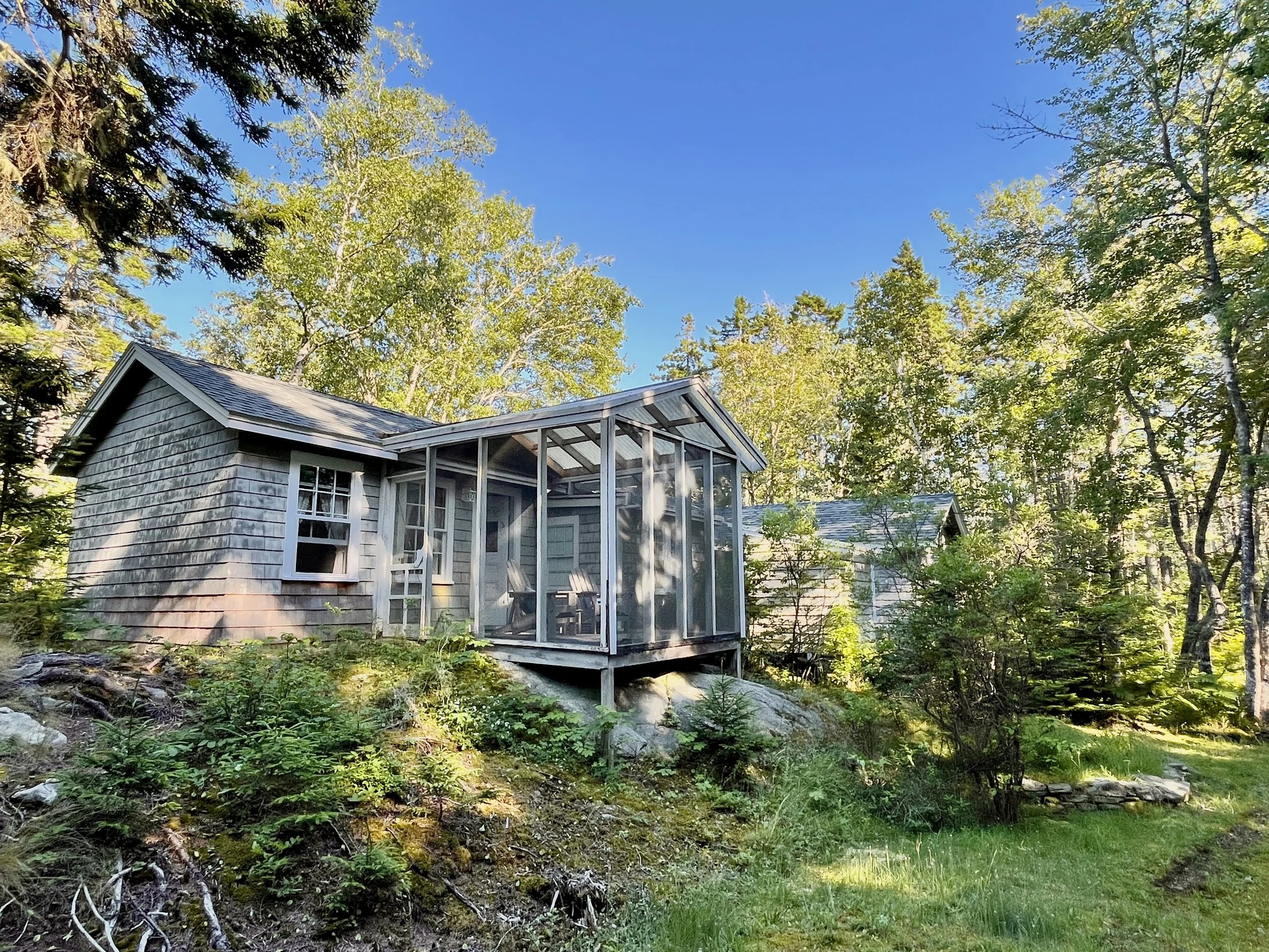 A small house with a screened-in porch surrounded by trees and greenery, located on a hill with rocks, plants, and a lawn in the foreground.