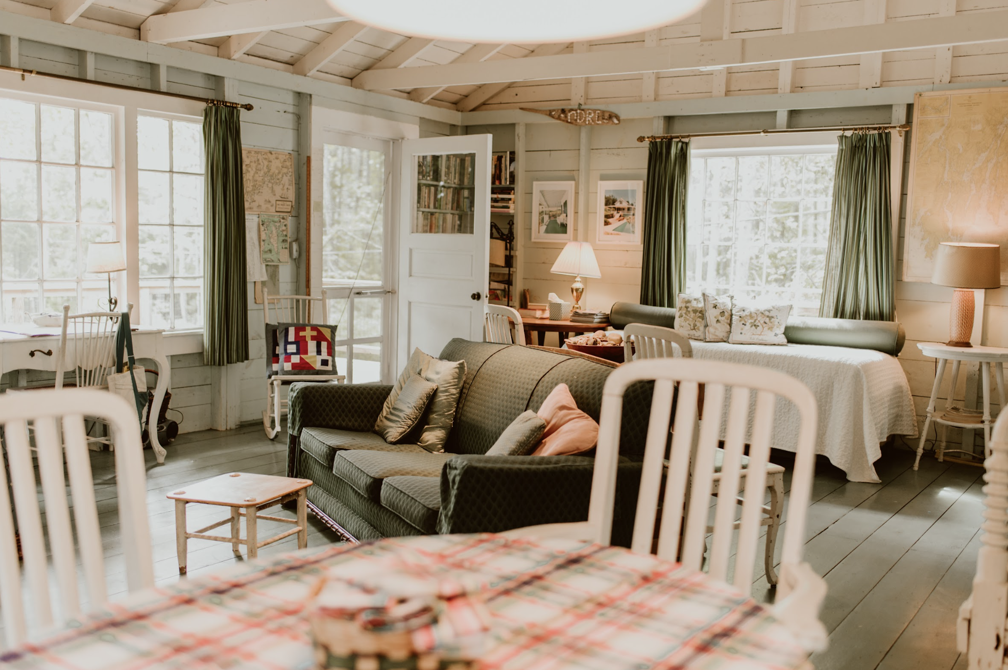 Interior of a cozy living room with large windows, a green sofa with pillows, a bed with white bedding, wooden chairs, and a round dining table with a red and white checkered tablecloth.