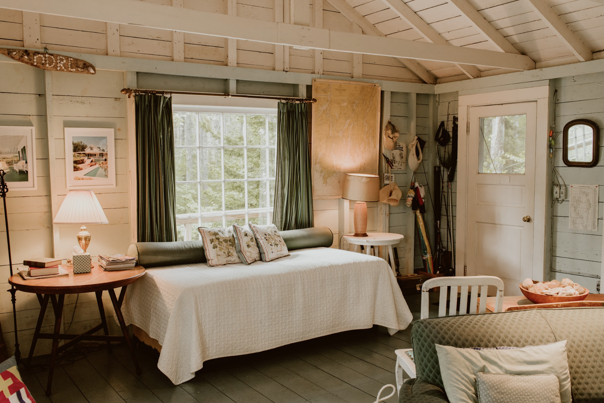 Cozy rustic interior of a small bedroom with a large window, bed with decorative pillows, side tables with lamps, and wall decorations including photographs and fishing gear.