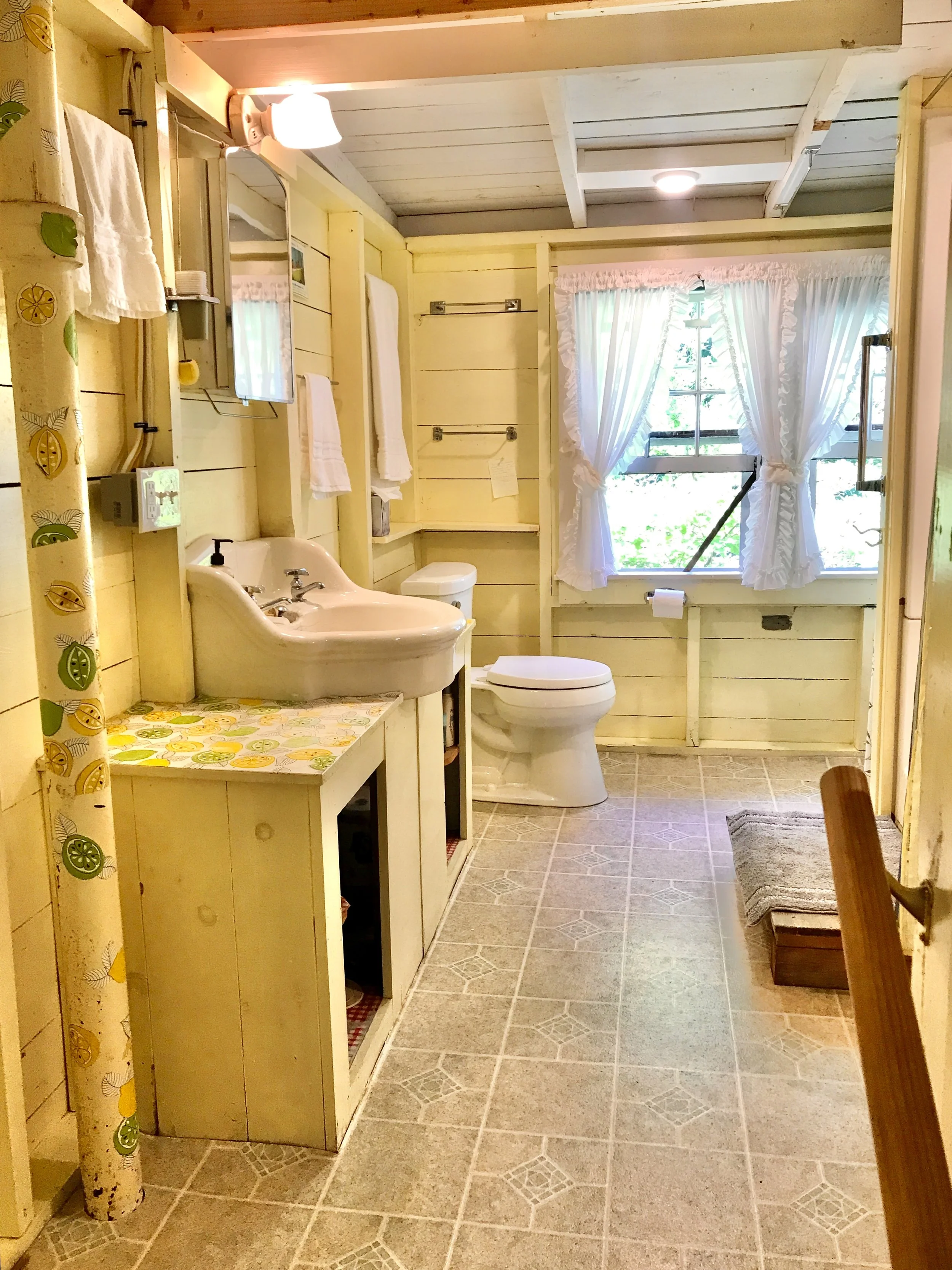 A rustic bathroom with yellow wooden walls, a small white sink, a toilet, and a window with white curtains. The floor is laminate, and a vertical pipe with a lemon pattern is visible on the left.