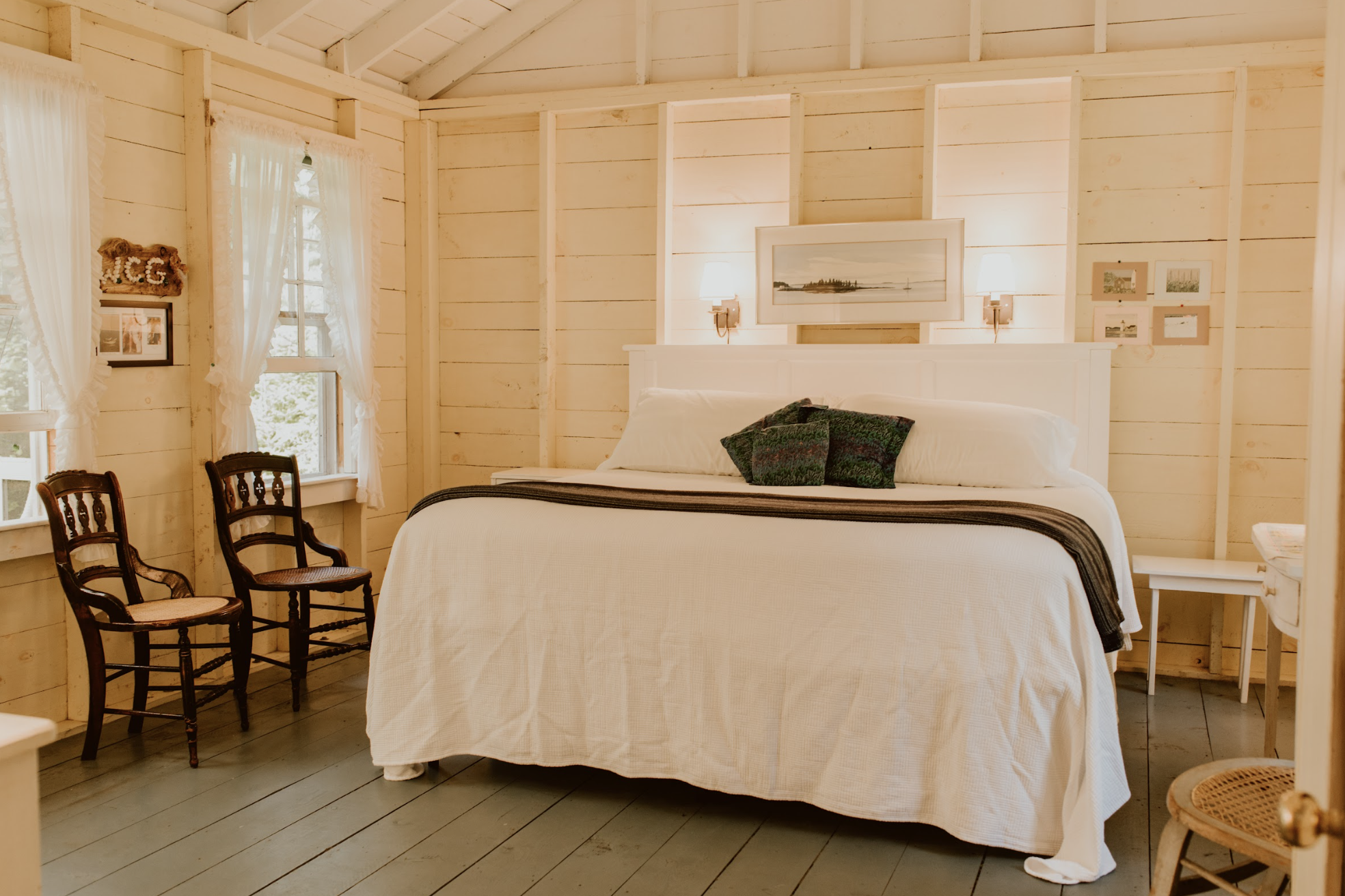 A bedroom with light-colored wooden walls, a large bed with white bedding, and two picture frames and a painting above the headboard. There are two small lamps on each side of the bed, two wooden chairs with woven seats near the window, and a small w