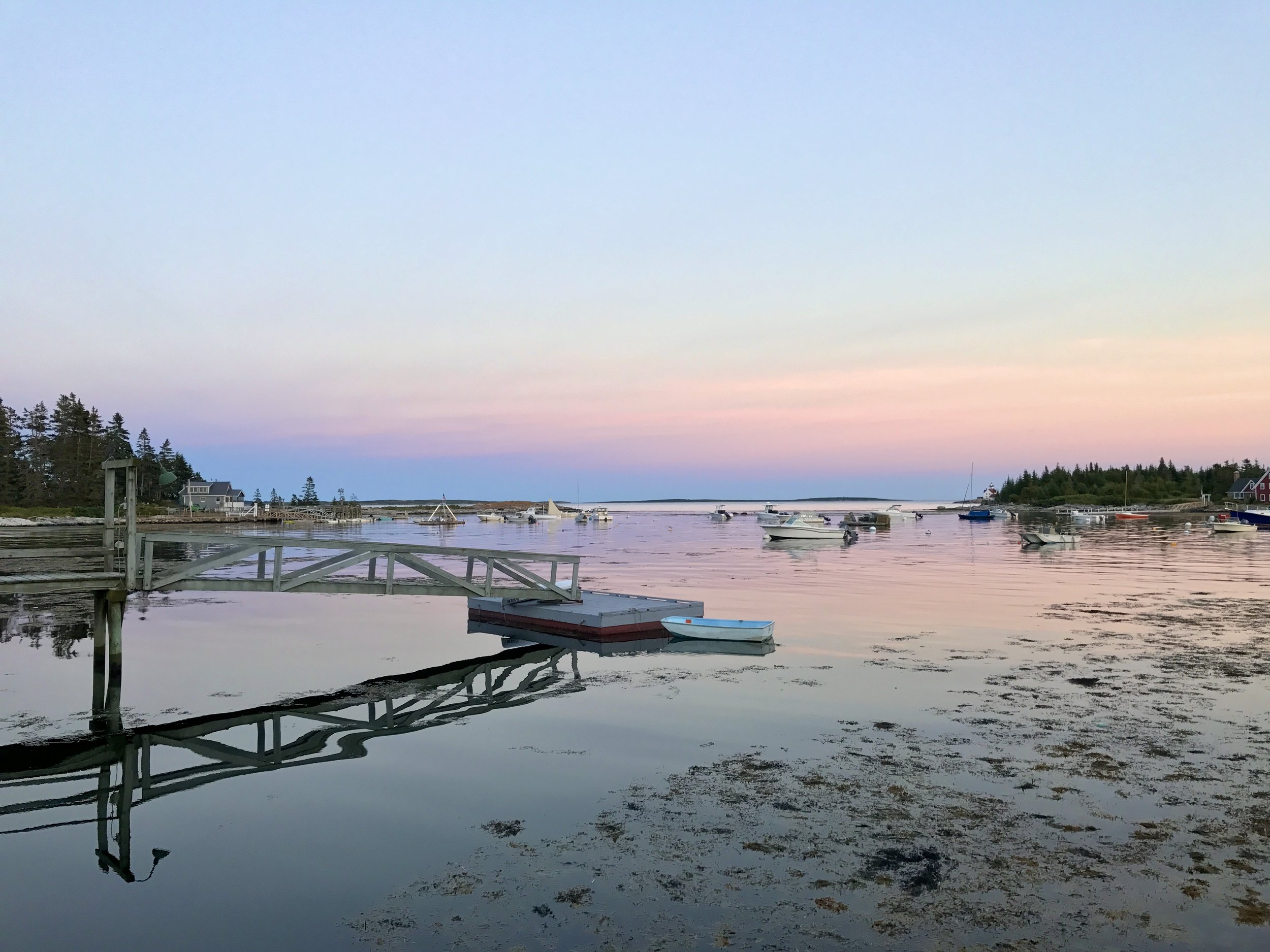 Calm harbor with boats anchored, wooden dock, and houses on wooded shoreline at sunset with pastel sky.