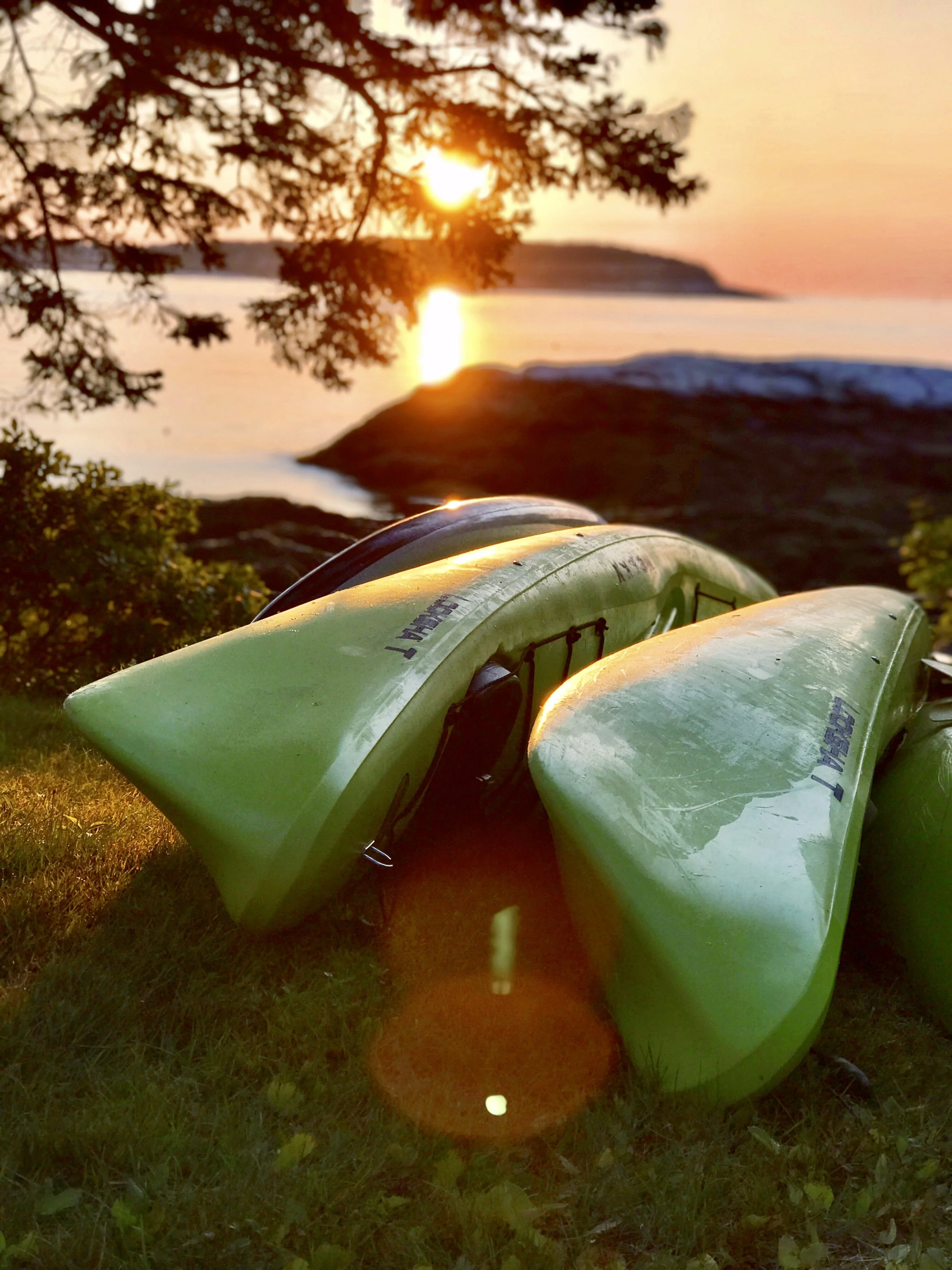 Two green kayaks resting on grass near a rocky shoreline at sunrise, with the sun rising over water and trees in the background.