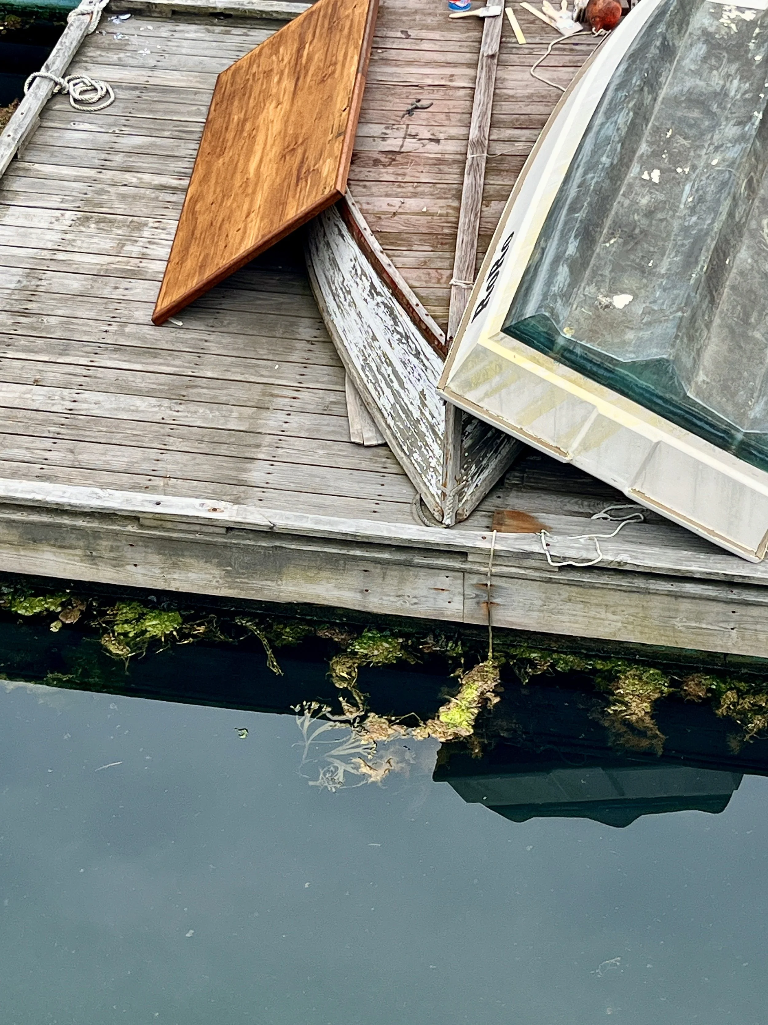 View of a dock with weathered wooden planks, a small wooden table, and an old boat with peeling paint, beside water with algae and plant debris along the edge.
