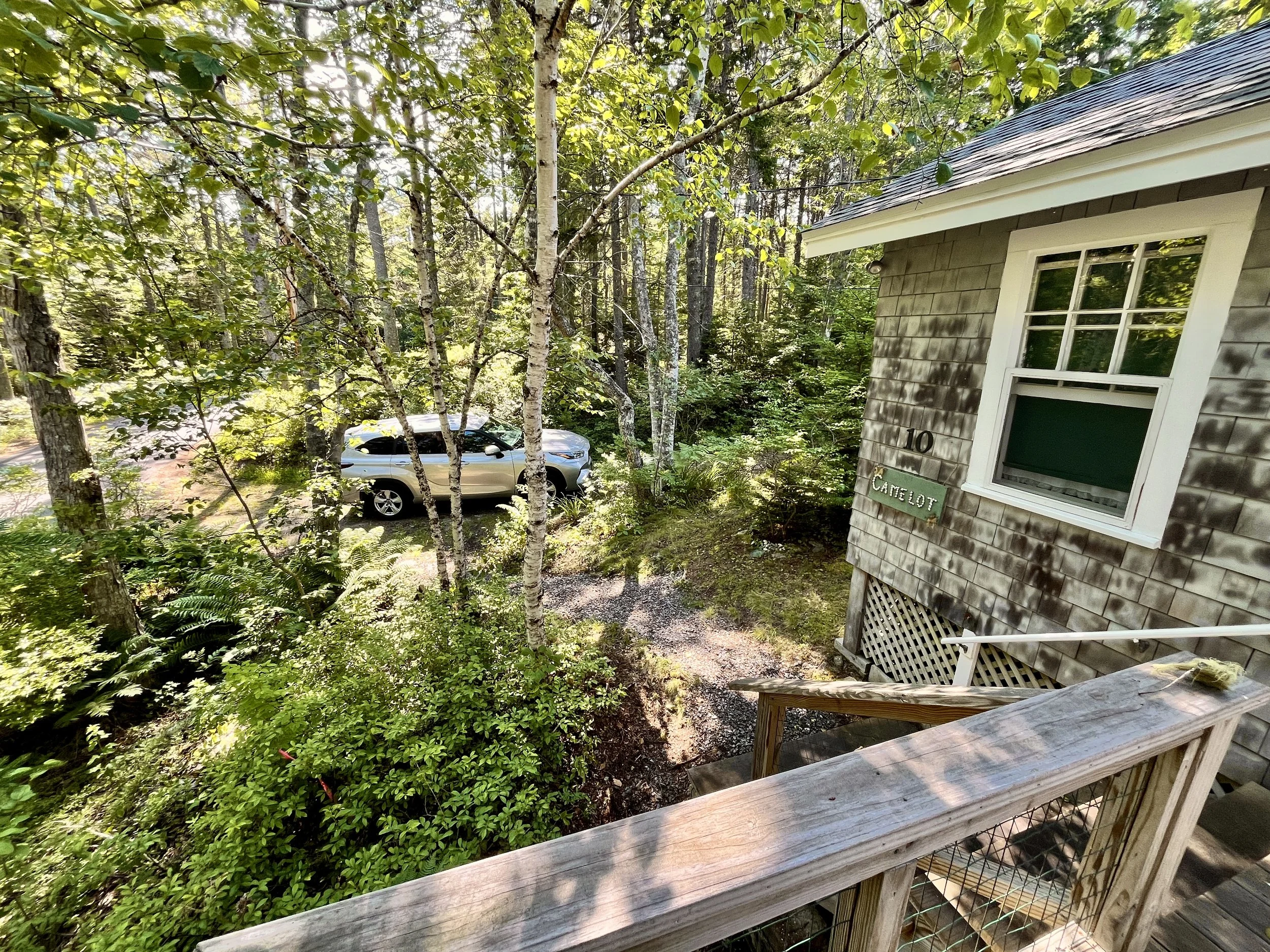 View of a wooded driveway and front walkway with a parked silver car, a small wooden porch, and a house with weathered wooden siding and a window.